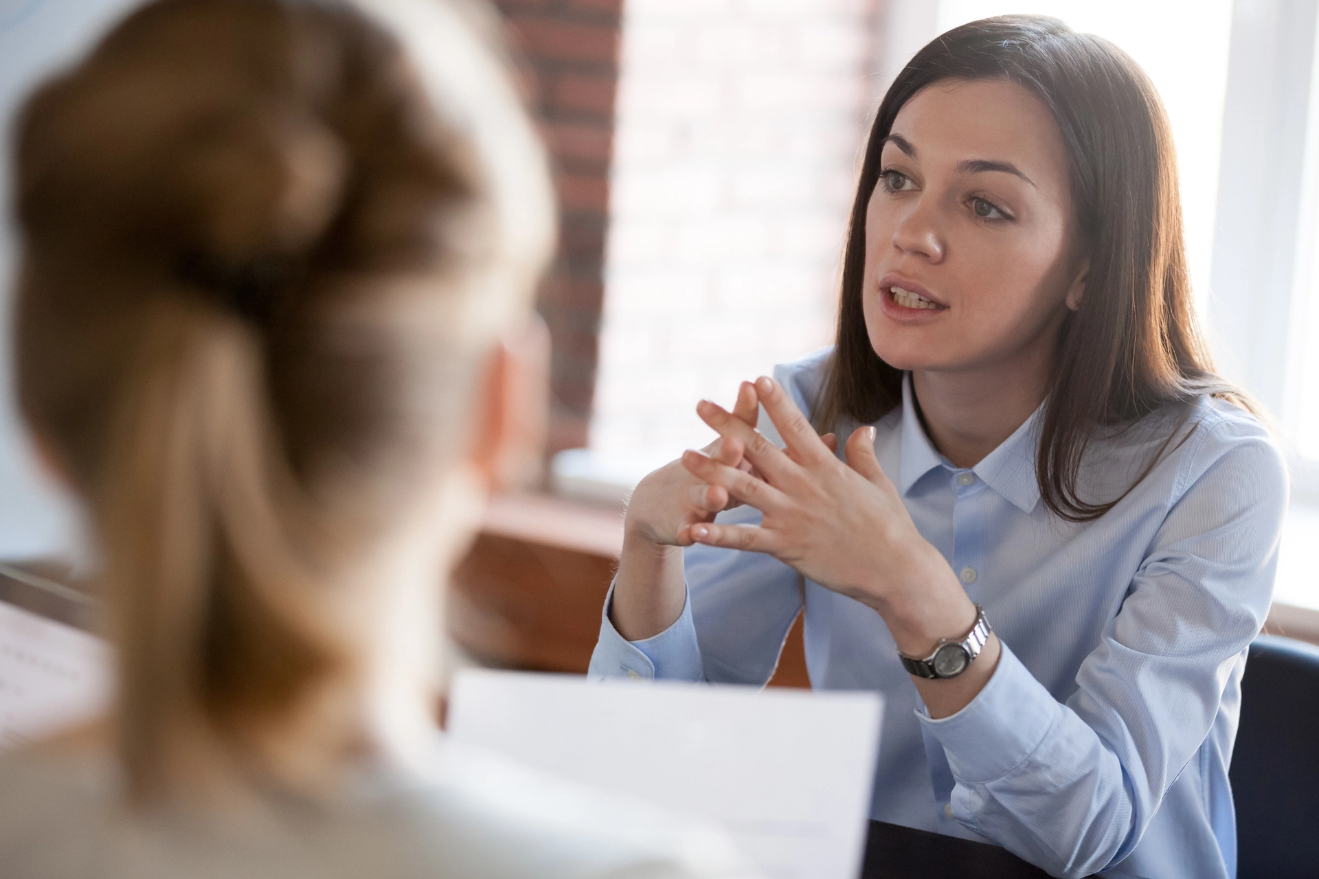 A woman in a blue shirt speaks with clasped hands across a table to another person.