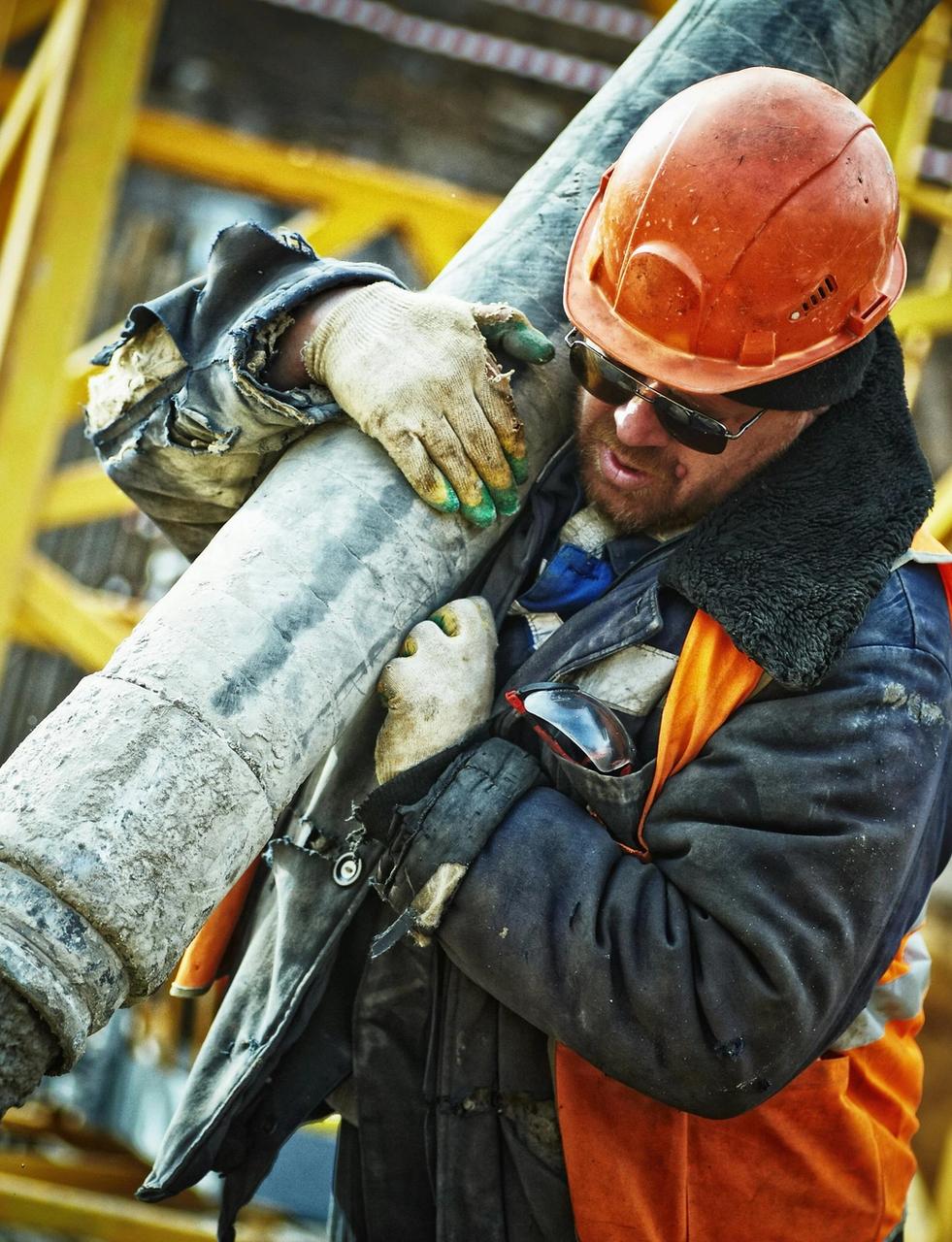 Construction worker in a hard hat and protective gear carrying a concrete pump hose as concrete flows out.