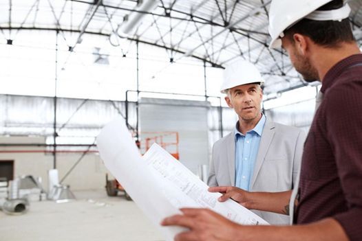 two men in hard hats are looking at a blueprint in a warehouse .