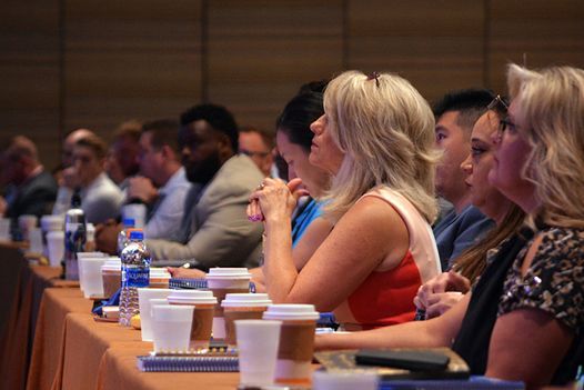 a group of people are sitting at a long table with coffee cups and water bottles .