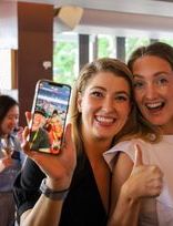 two women are posing for a picture with a cell phone and giving a thumbs up .