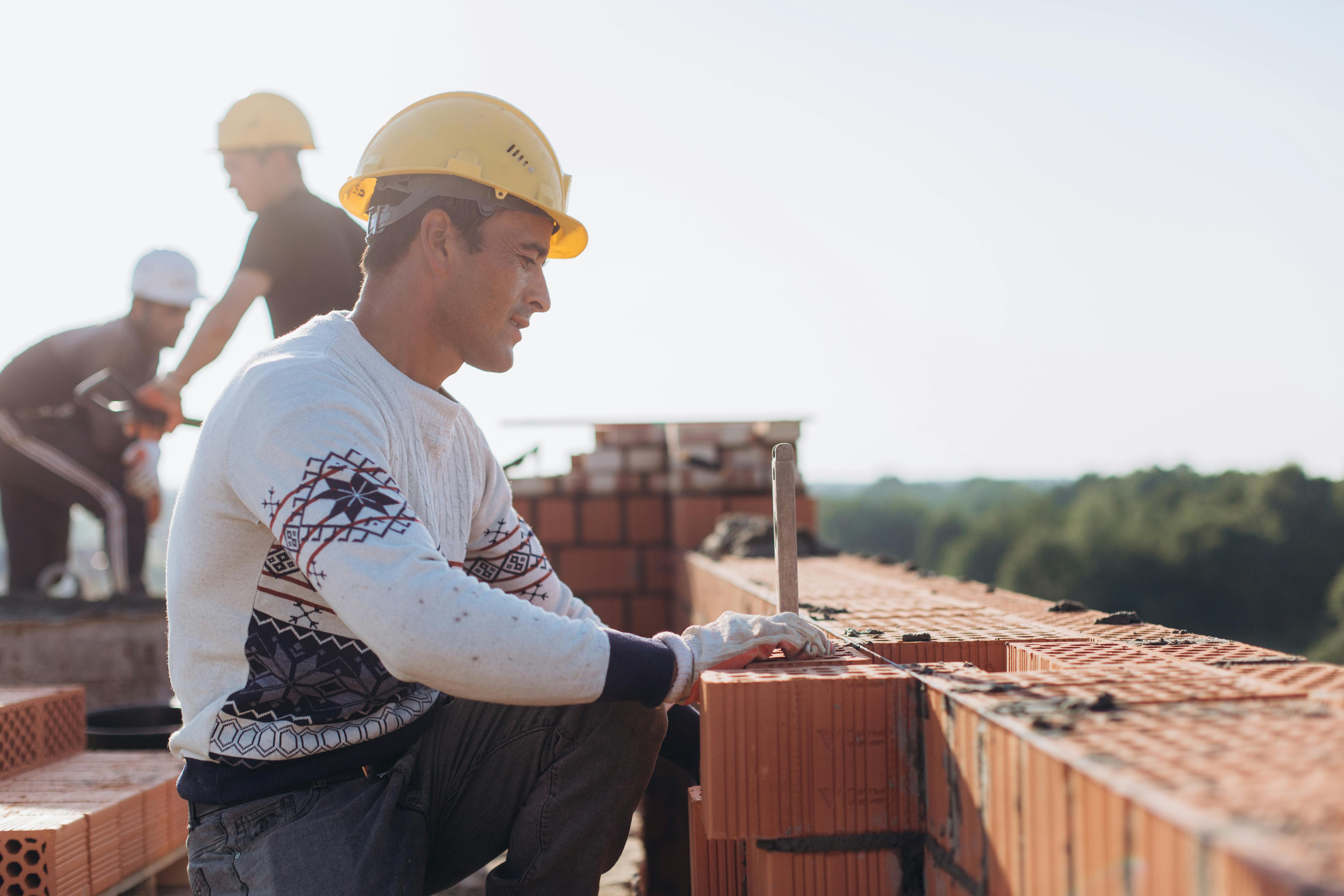 A construction worker in a yellow hard hat lays bricks on an outdoor site.