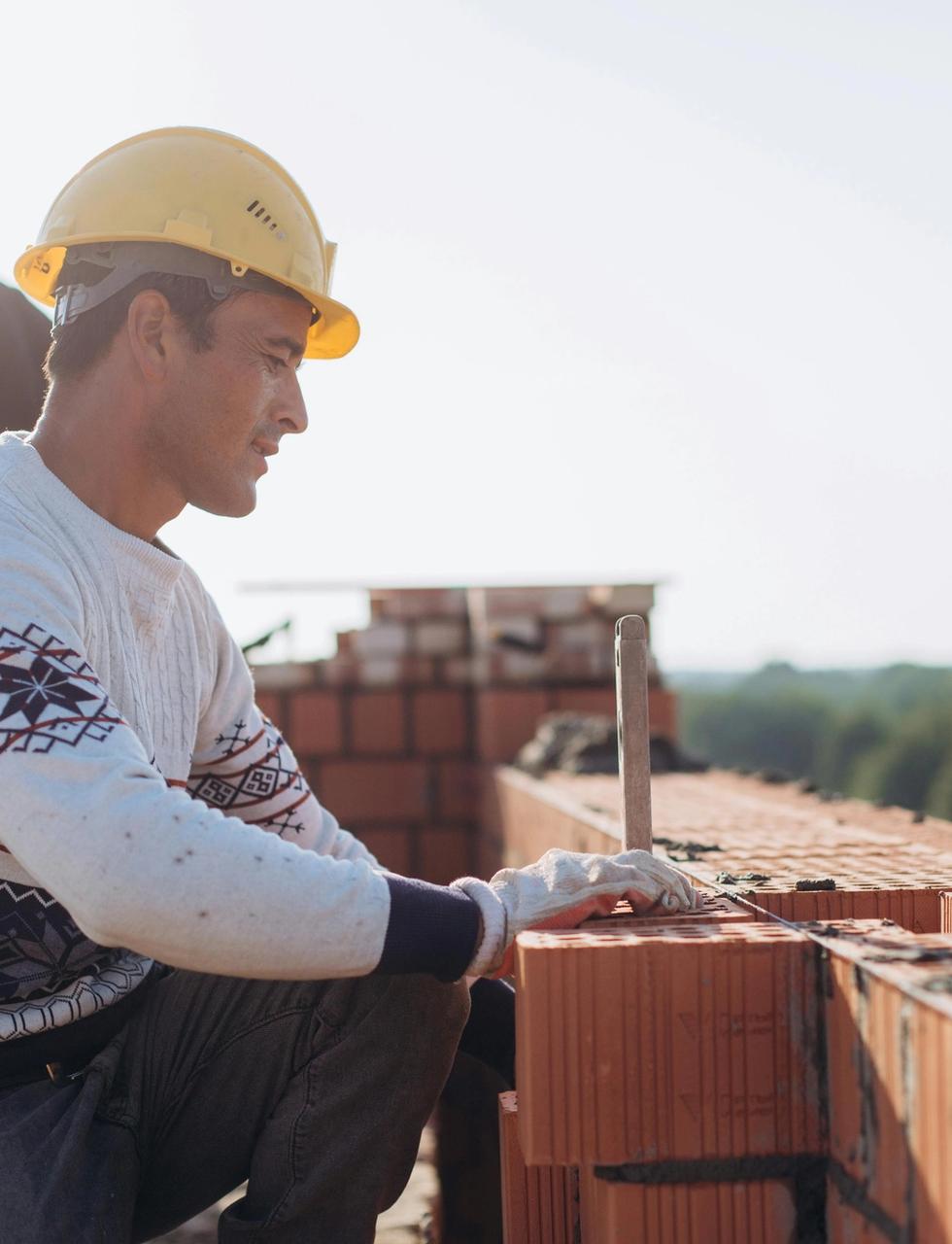 A construction worker in a yellow hard hat lays bricks on an outdoor site.