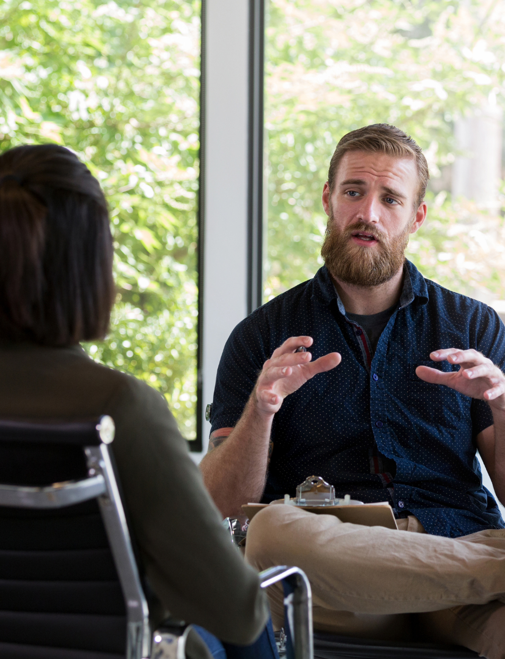A bearded man gestures while talking to a woman in an office setting.