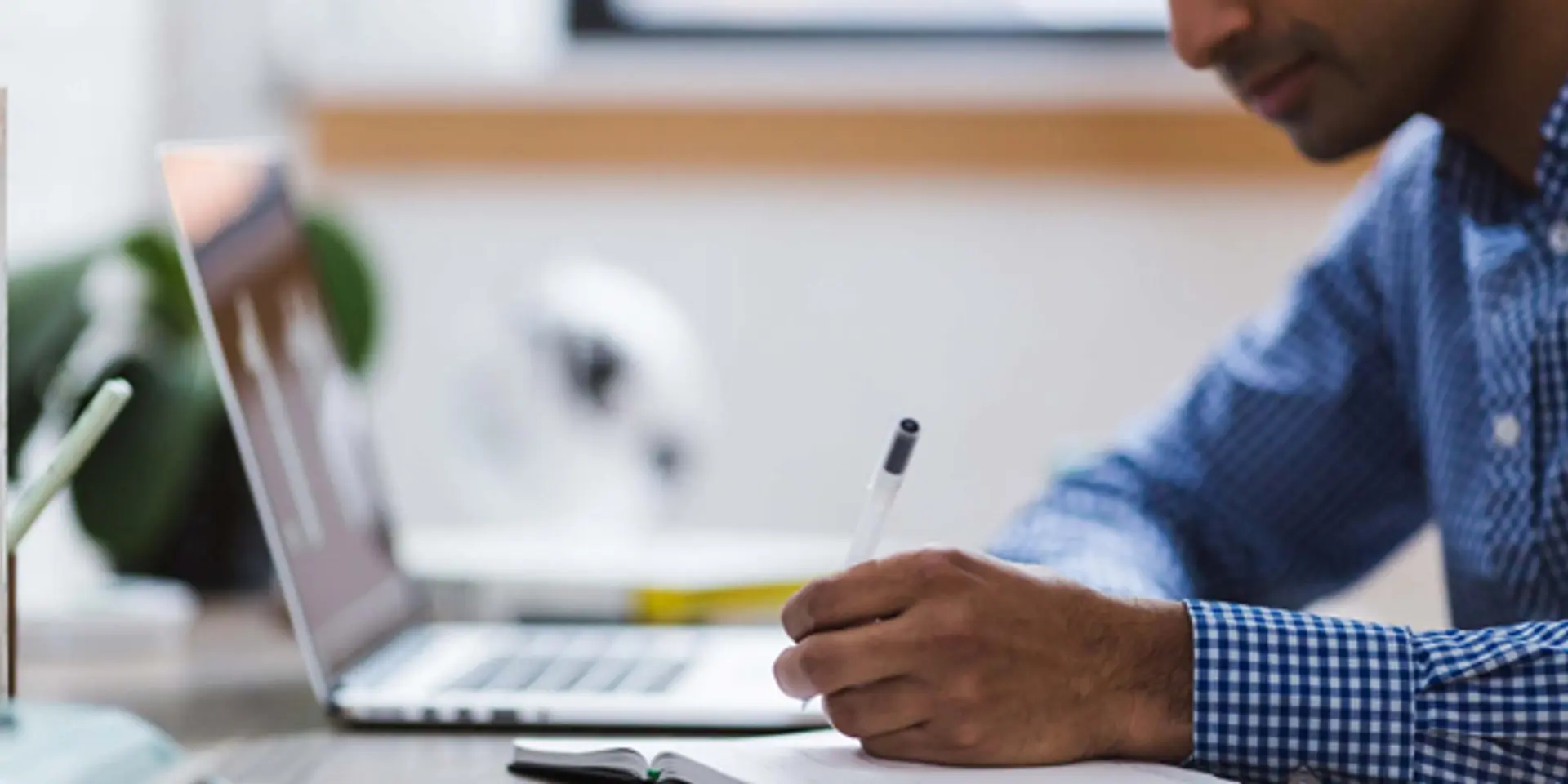 a man is sitting at a desk writing in a notebook in front of a laptop computer .