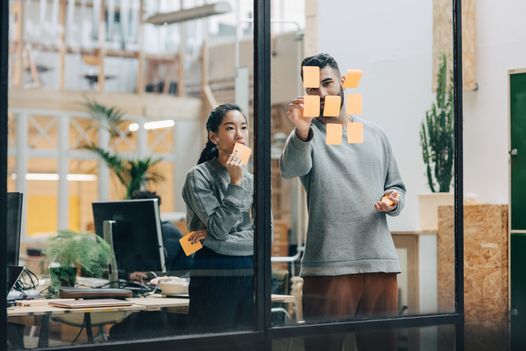 a man and a woman are looking at sticky notes on a glass wall .