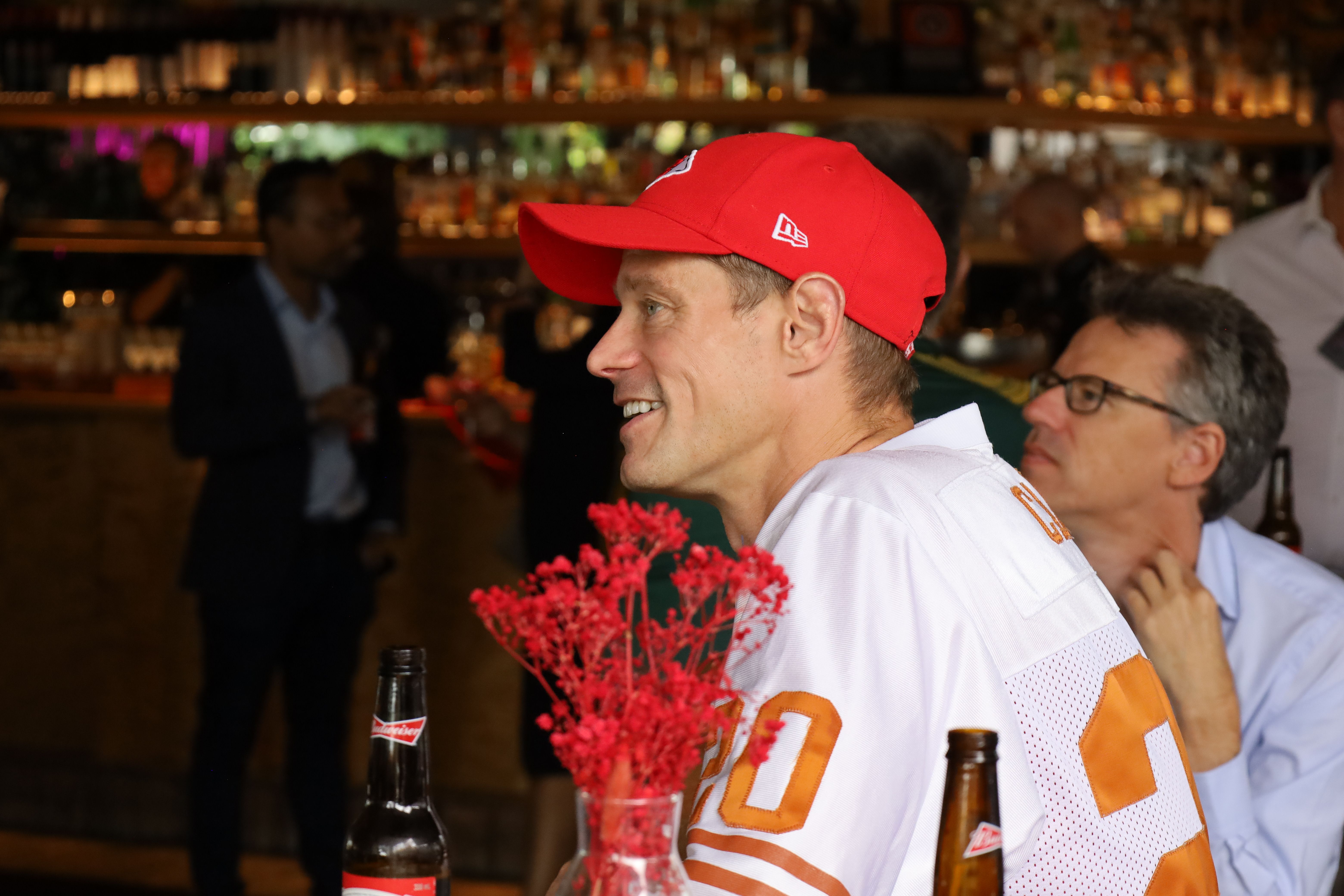 A man in a red cap and white jersey smiles in a bar setting.