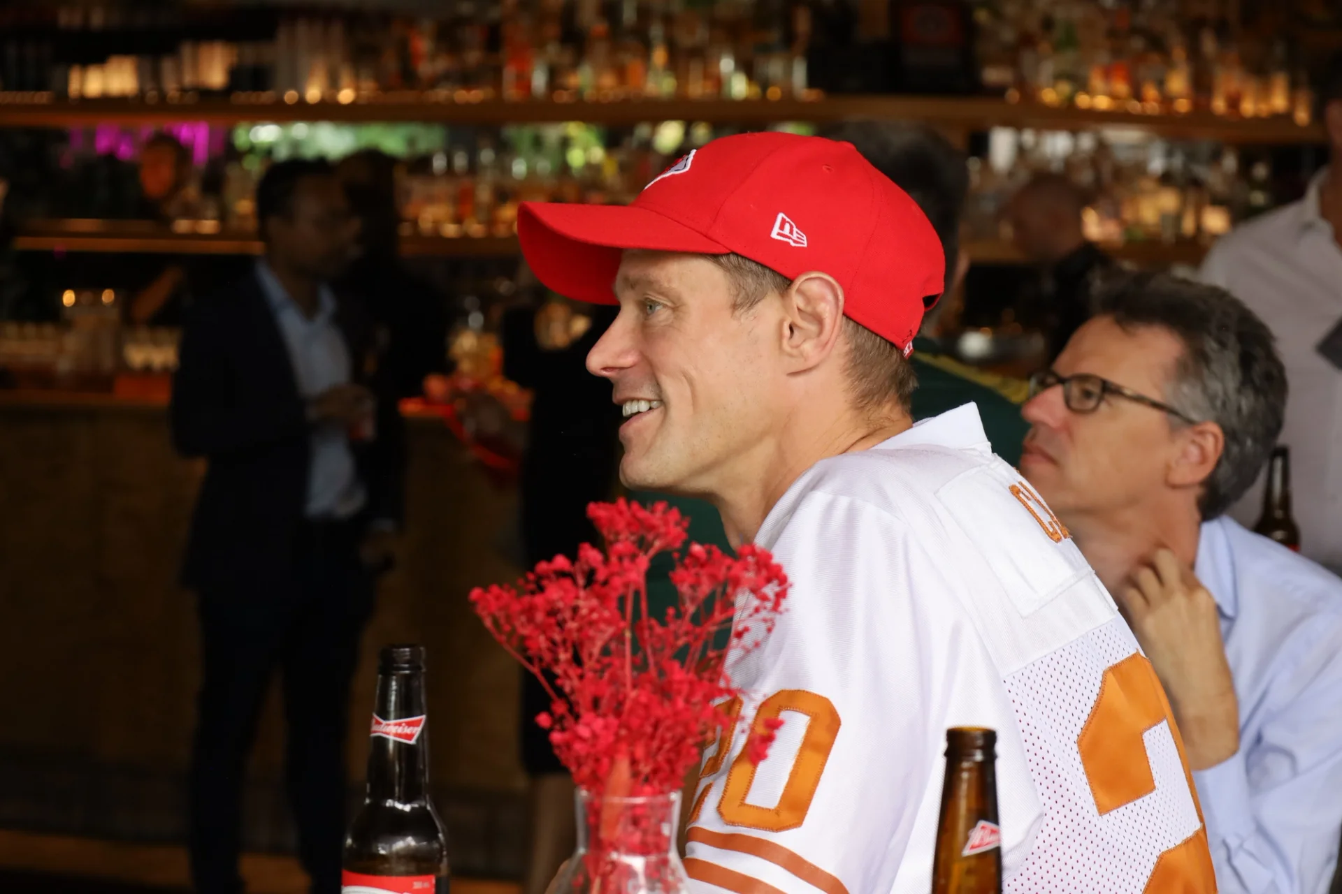 A man in a red cap and white jersey smiles in a bar setting.