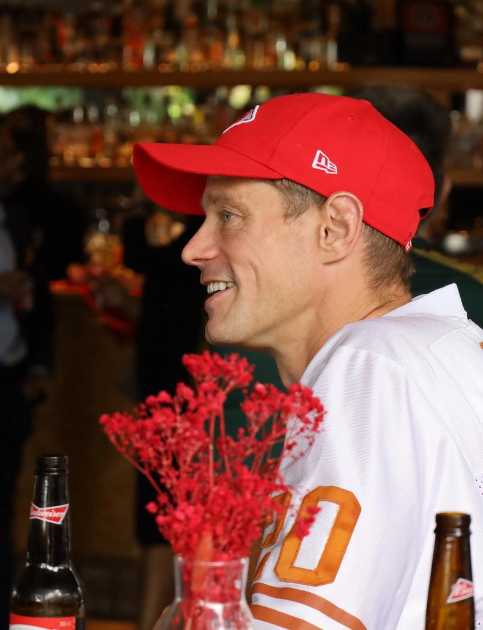 A man in a red cap and white jersey smiles in a bar setting.