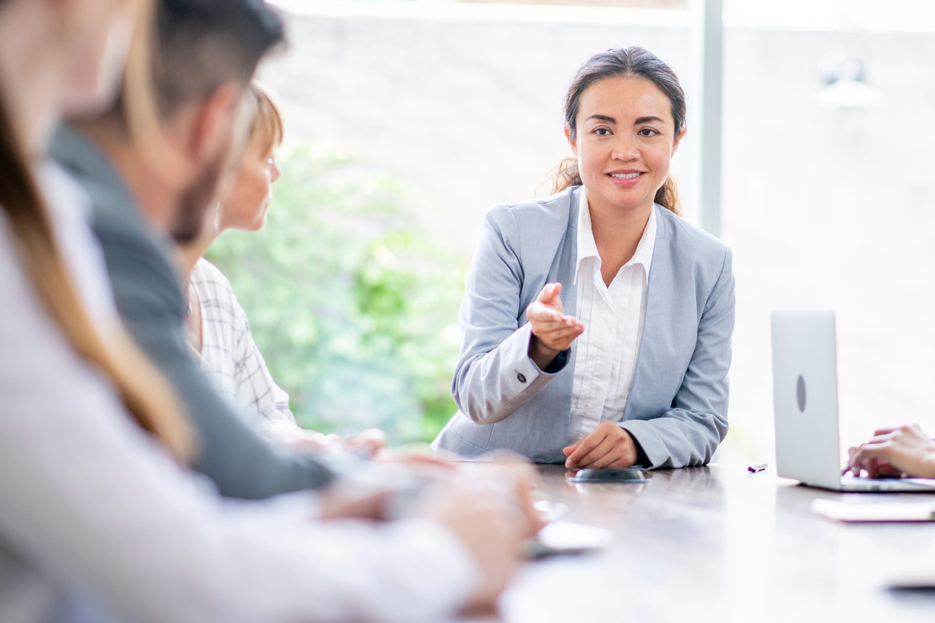 A smiling businesswoman in a suit gestures while speaking at a meeting.