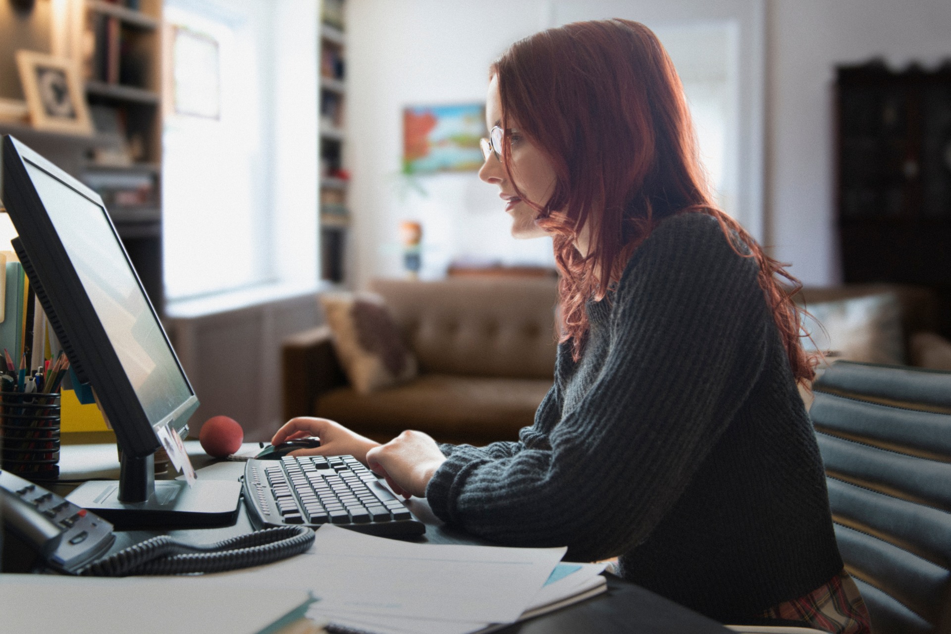 A red-haired woman with glasses sits at a desk, working on a desktop computer.