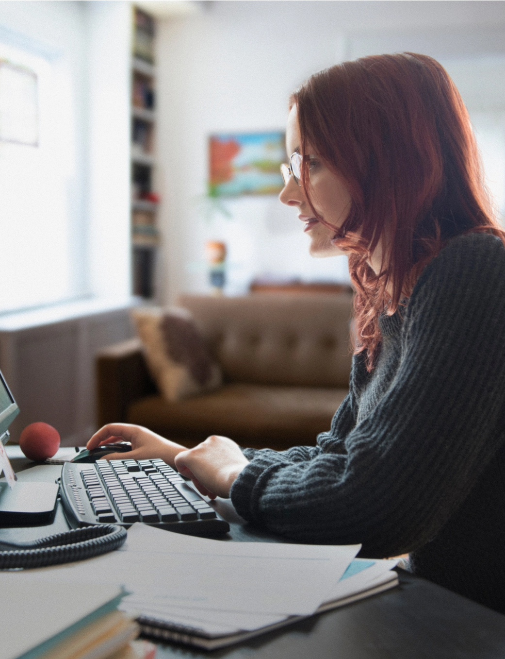 A woman with red hair and glasses works on a desktop computer at home.