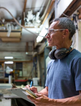 Man in safety glasses and headphones writing on a clipboard in a workshop.