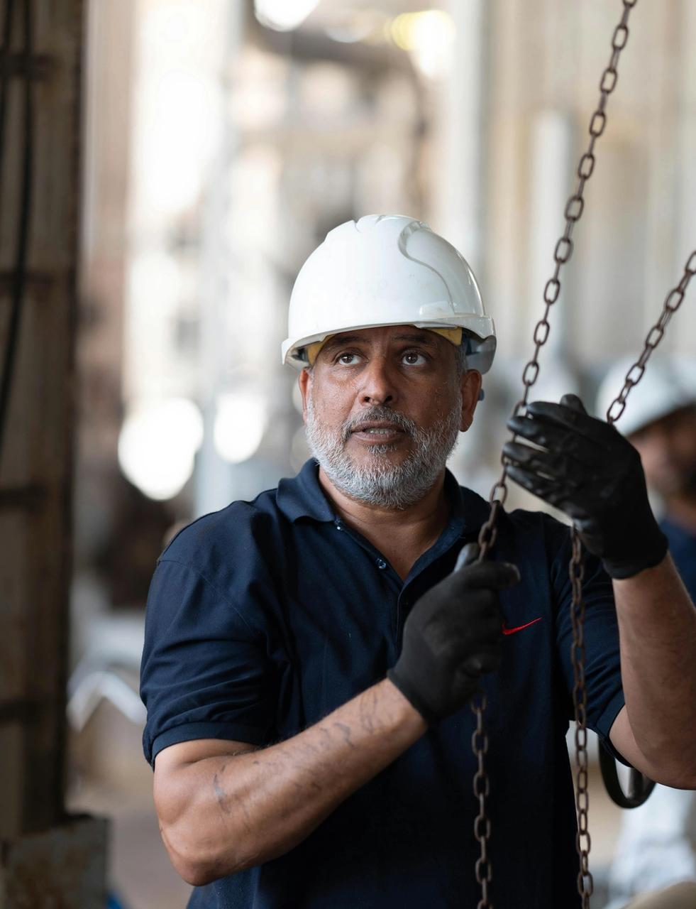 A bearded man in a white hard hat and work gloves holds chains, looking up in an industrial facility.