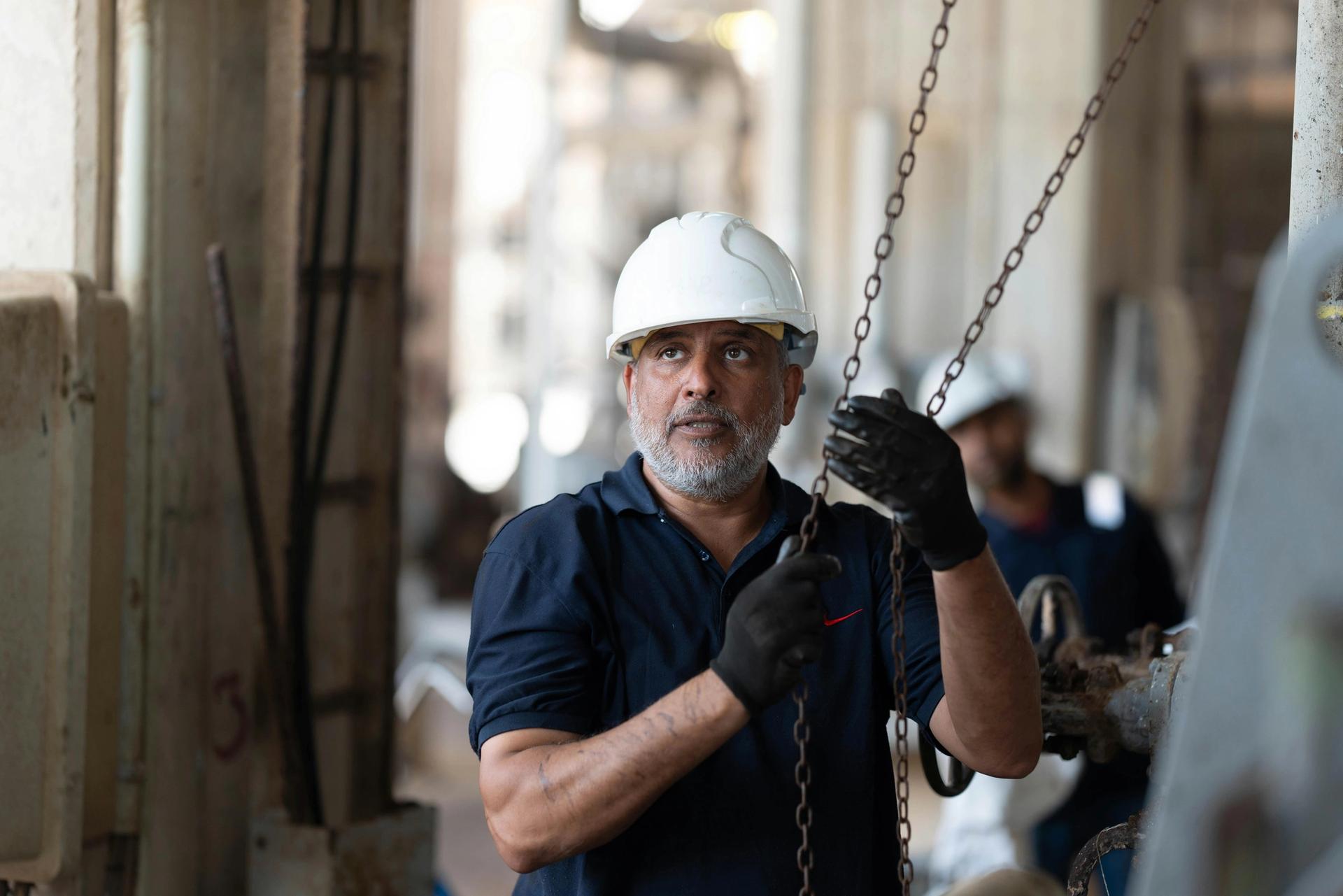 A bearded man in a white hard hat and work gloves holds chains, looking up in an industrial facility.