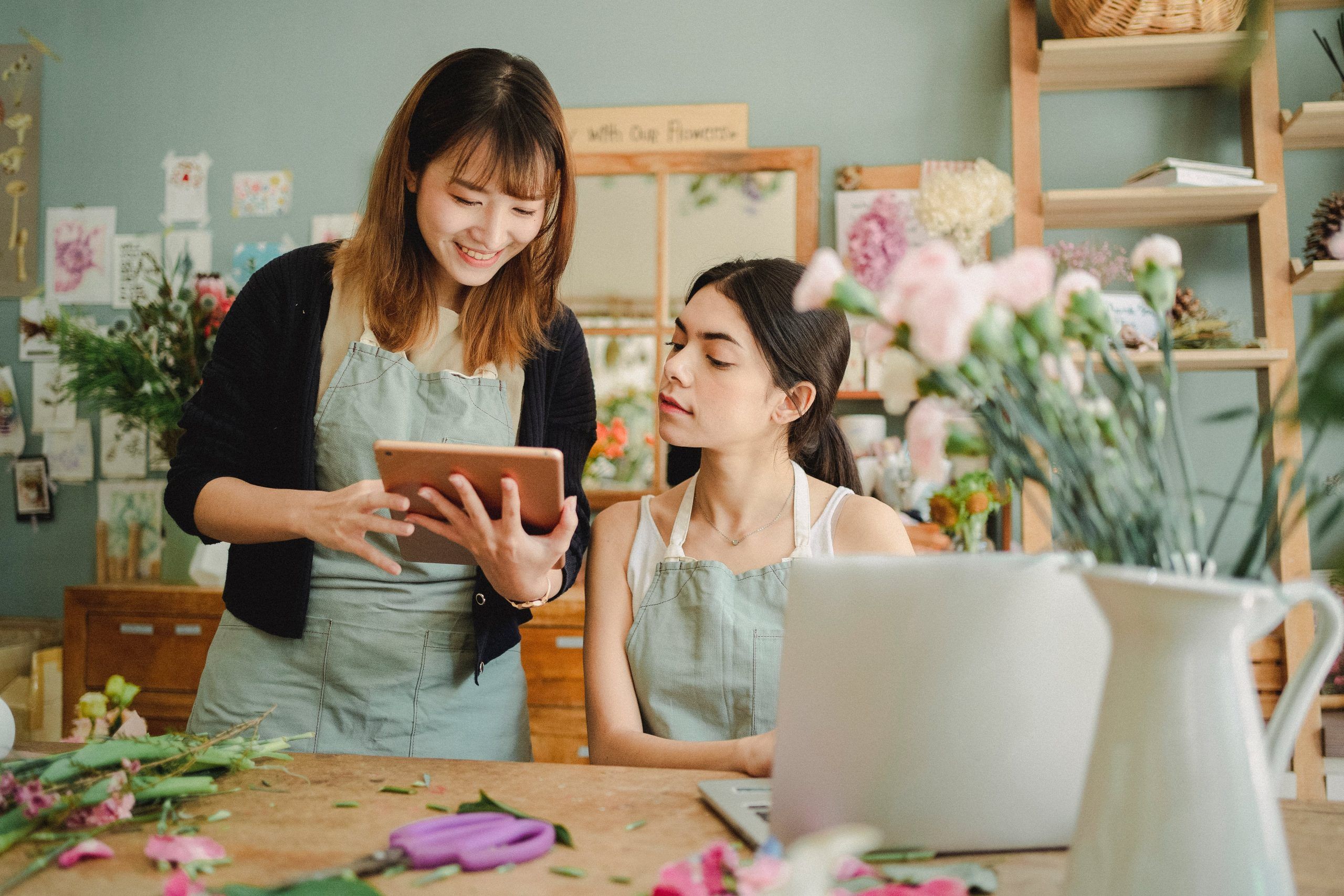 two women are sitting at a table in a flower shop looking at a tablet .