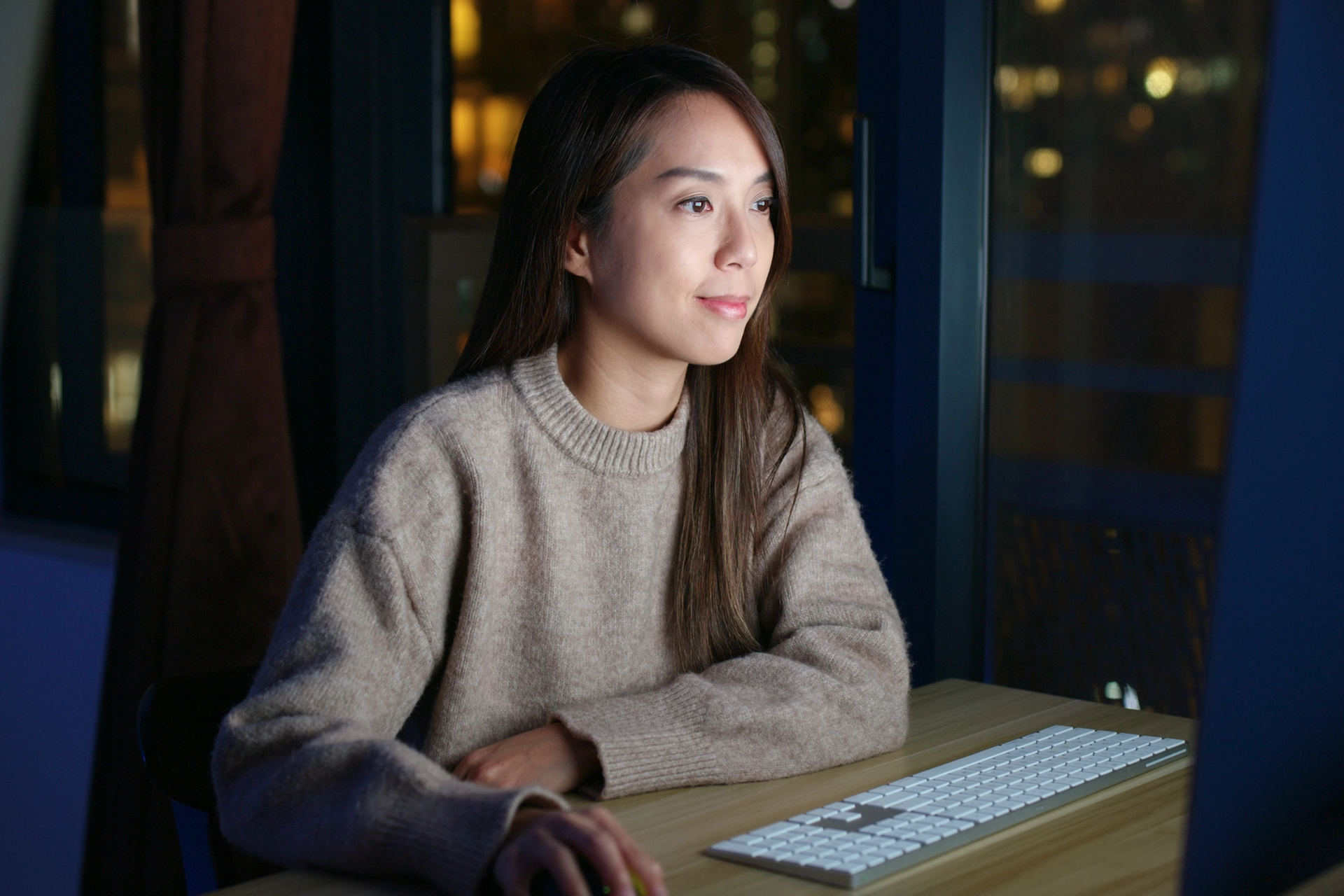 A woman works at a computer at night, with city lights in the background.