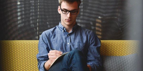 a young man is sitting on a couch writing in a notebook .