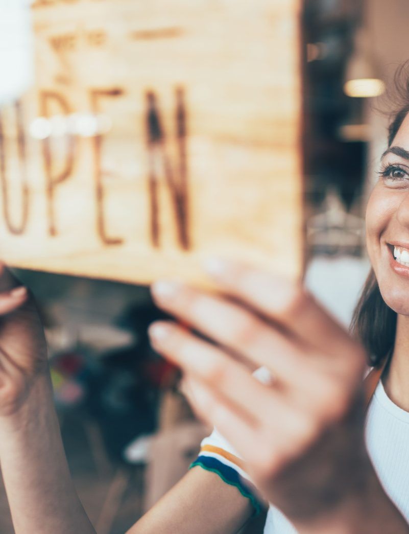 A smiling woman hangs an "OPEN" sign on a store window.