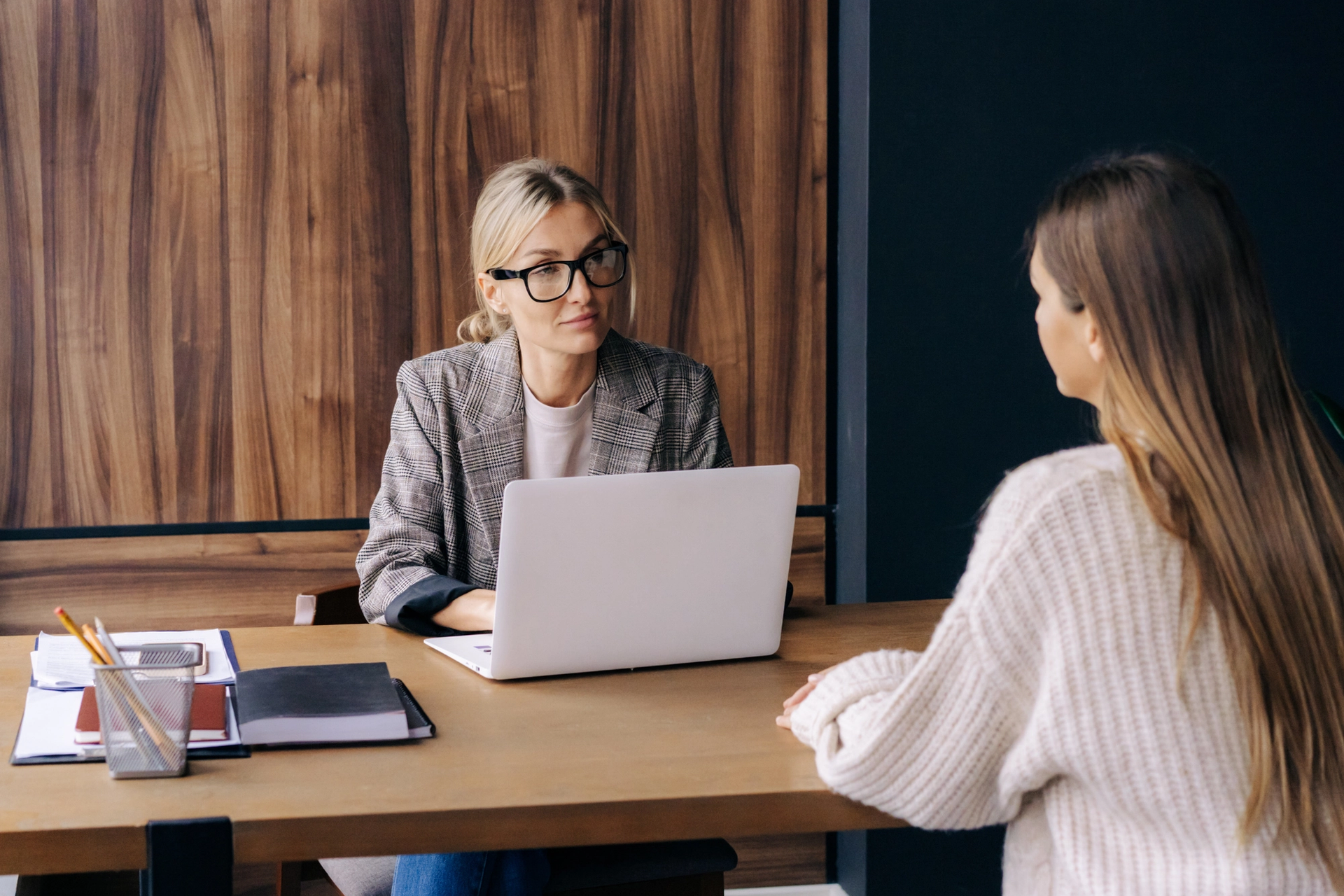 A woman with glasses at a desk with a laptop, interviewing another woman.