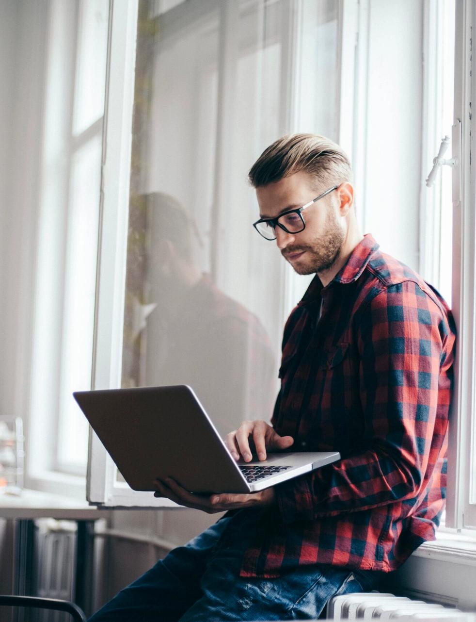 a man is sitting on a window sill using a laptop computer .