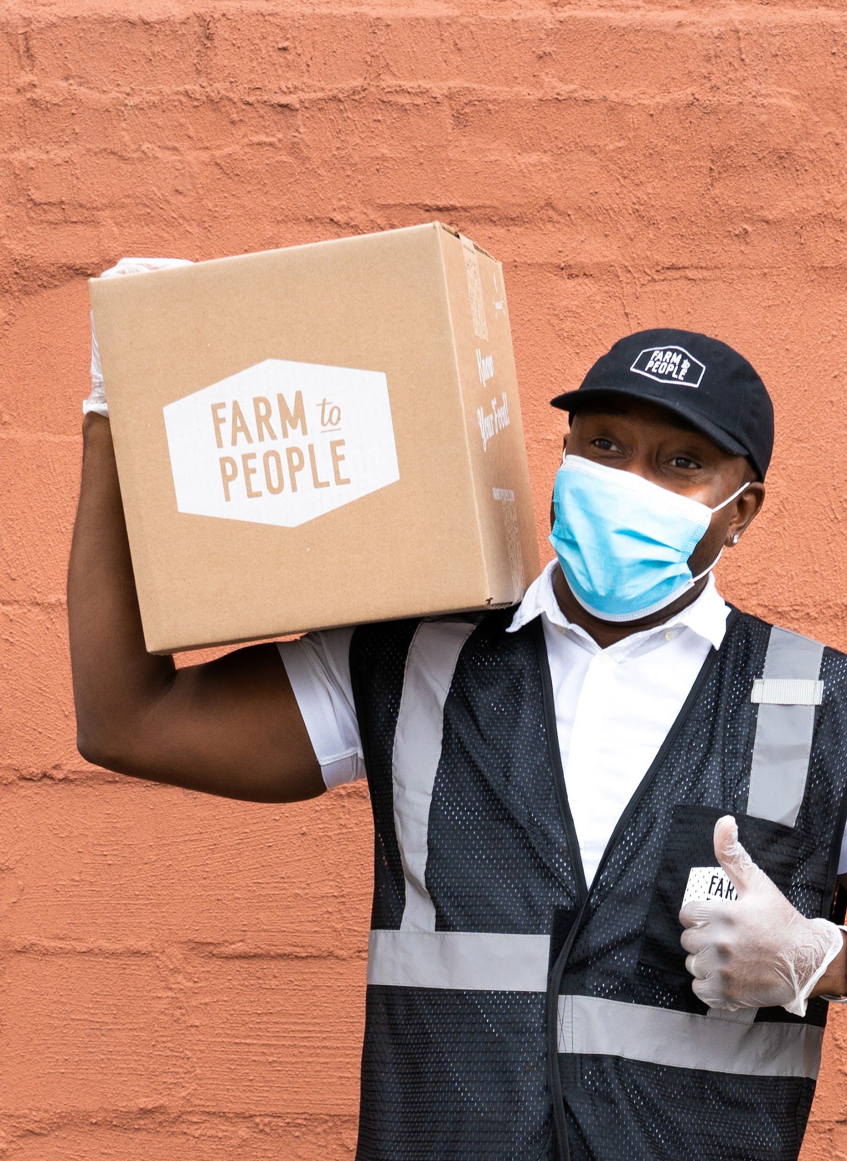 Deliver driver wearing reflective safety vest, mask and gloves while holding a branded Farm To People box on his shoulder