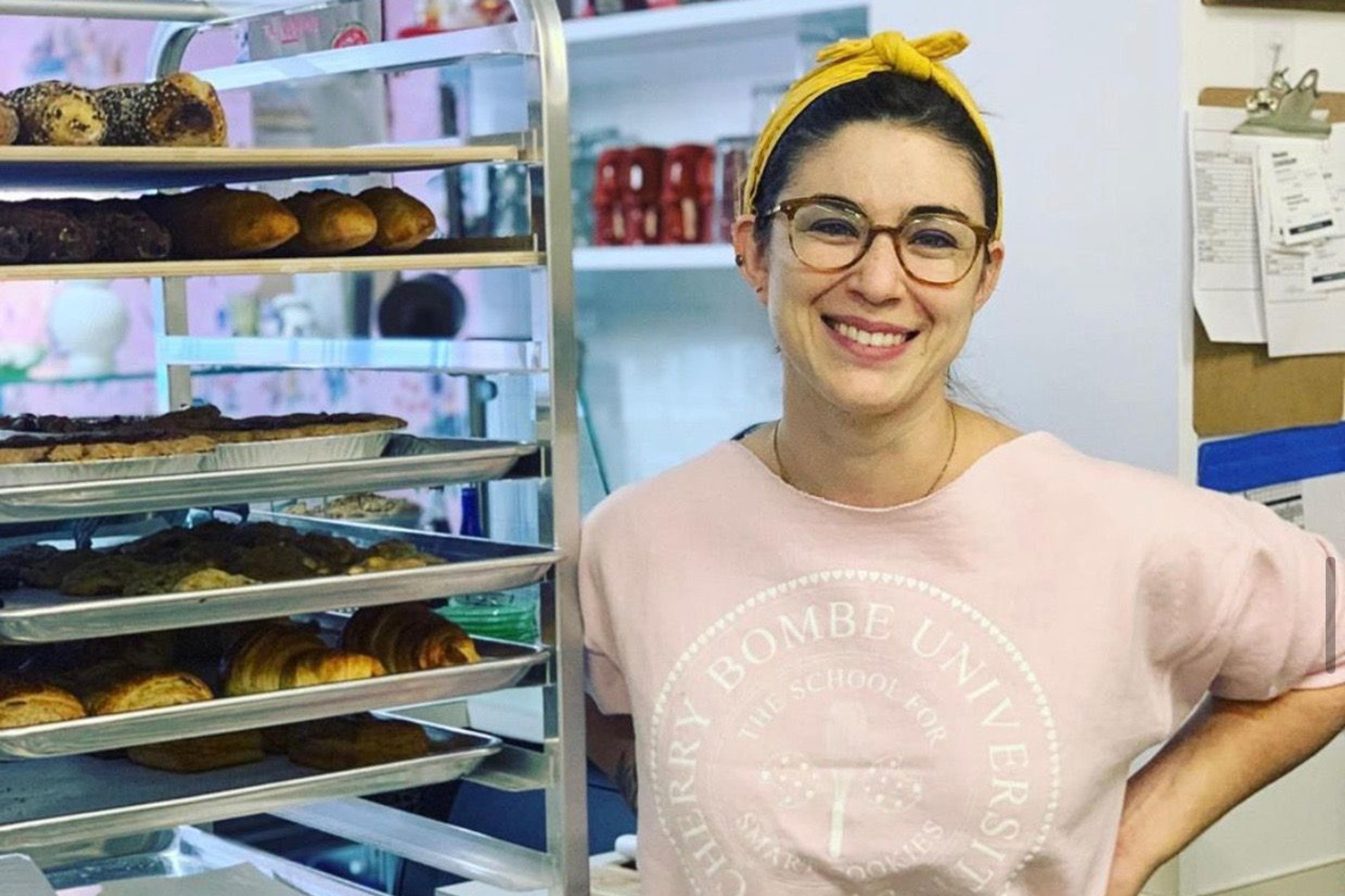 A woman wearing glasses and a headband stands next to a bakery rack filled with pies and pastries