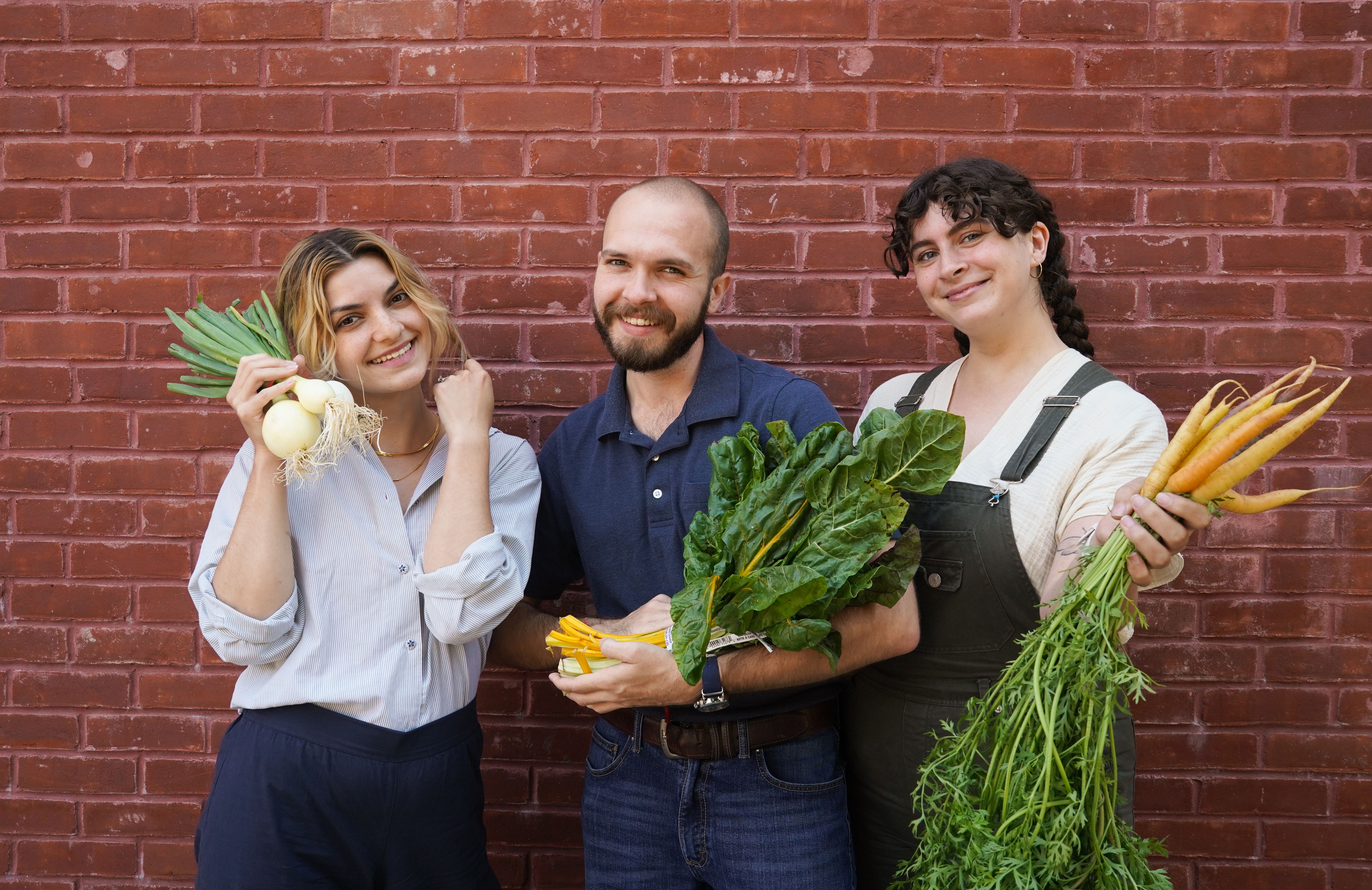 Three people pose smiling in front of a brick wall holding fresh vegtables.