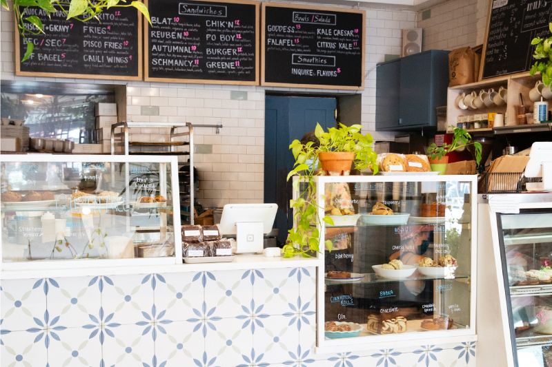 A tile faced cafe counter featuring bakery items in a display case in front of a chalk board menu