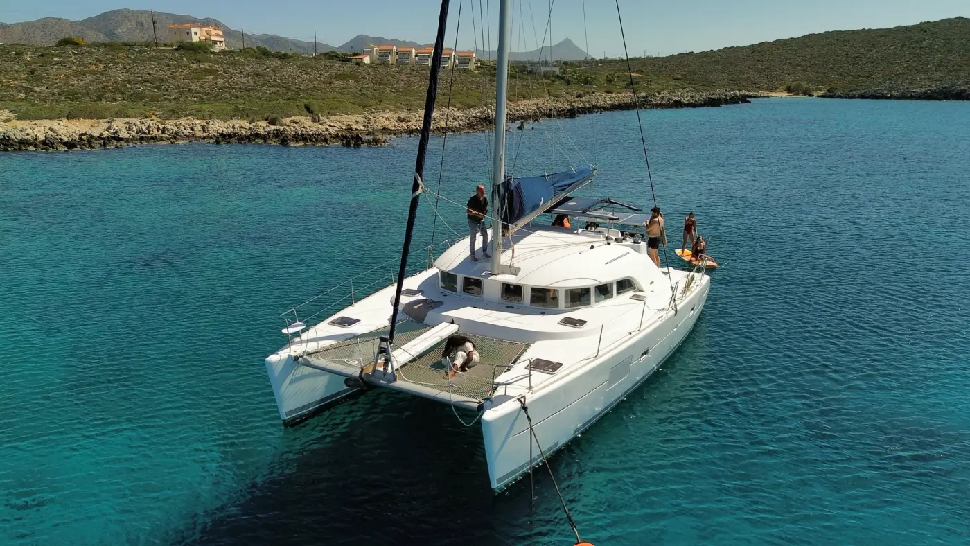 Crewed catamaran anchored in a calm, crystal-clear Greek bay with guests relaxing on the foredeck trampoline