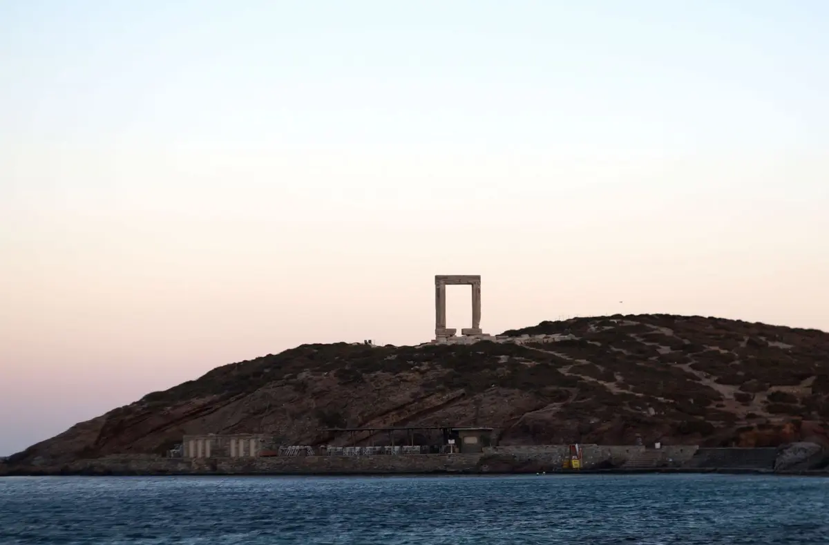 The Portara of Naxos at dusk seen from the water, the marble gate of an unfinished Temple of Apollo above the harbour.