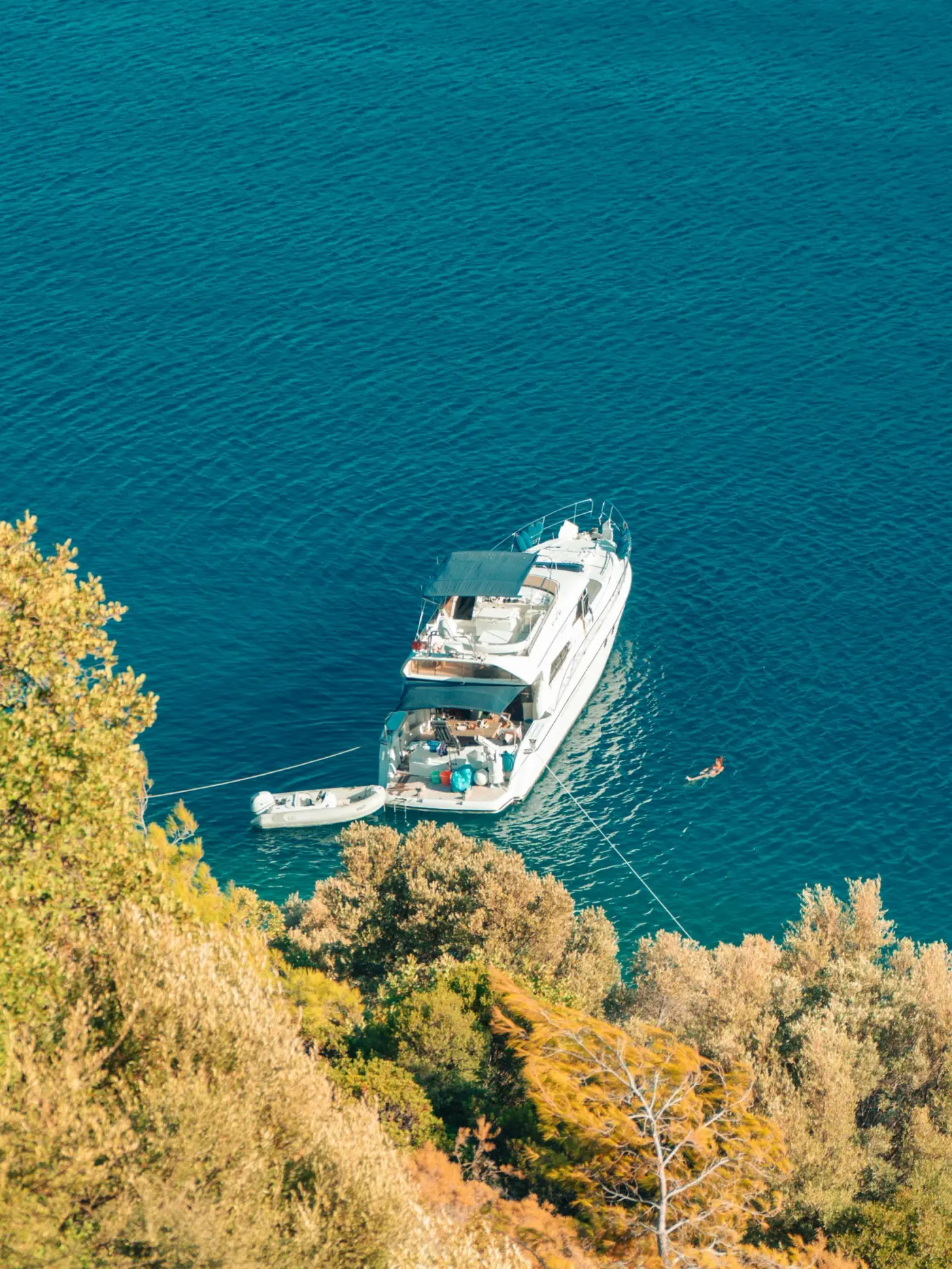 Motor yacht anchored in a sheltered Greek cove with a swimmer nearby, viewed from a hillside through Mediterranean greenery