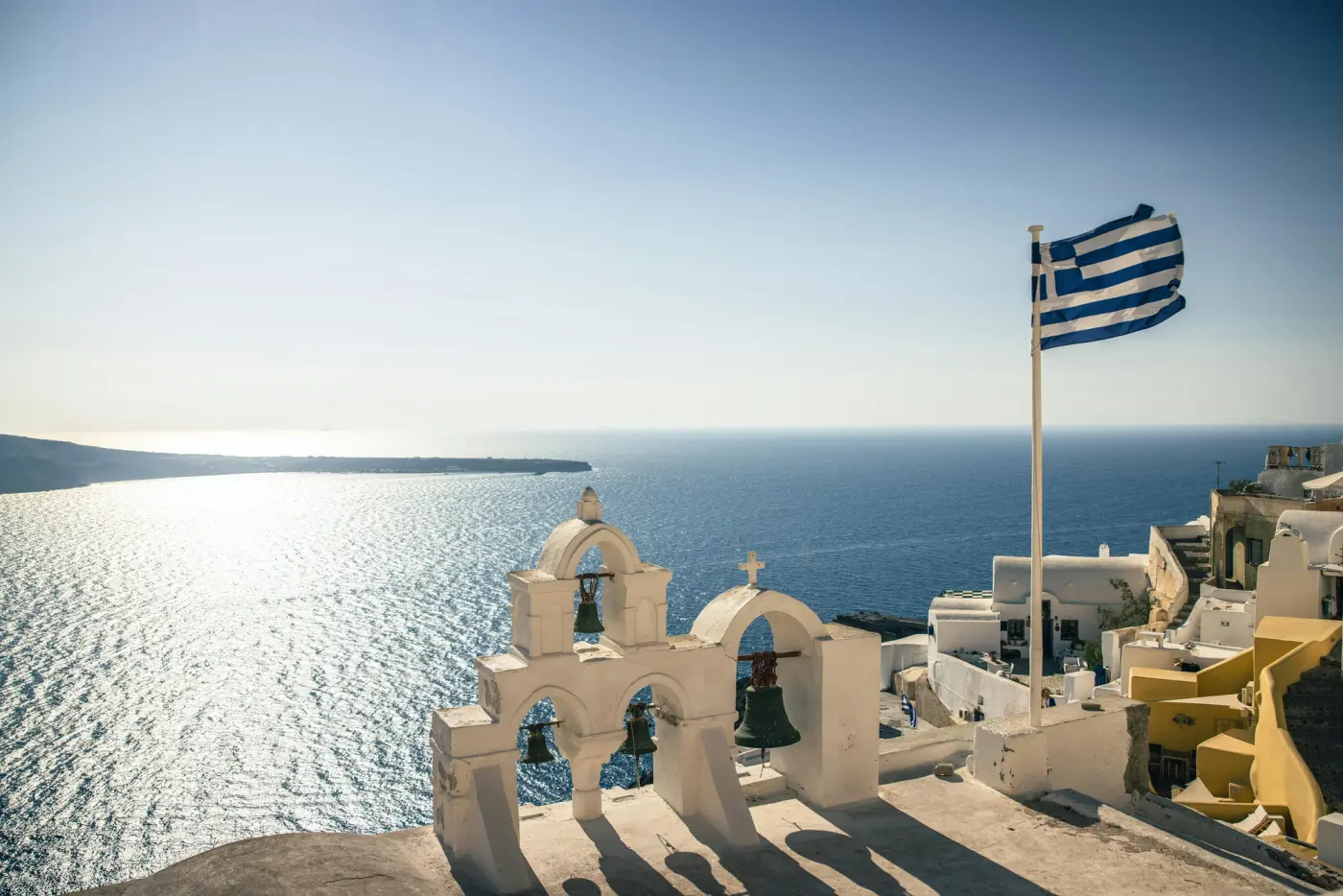 Greek flag waving beside a traditional white bell tower overlooking the Aegean Sea in Santorini, Greece