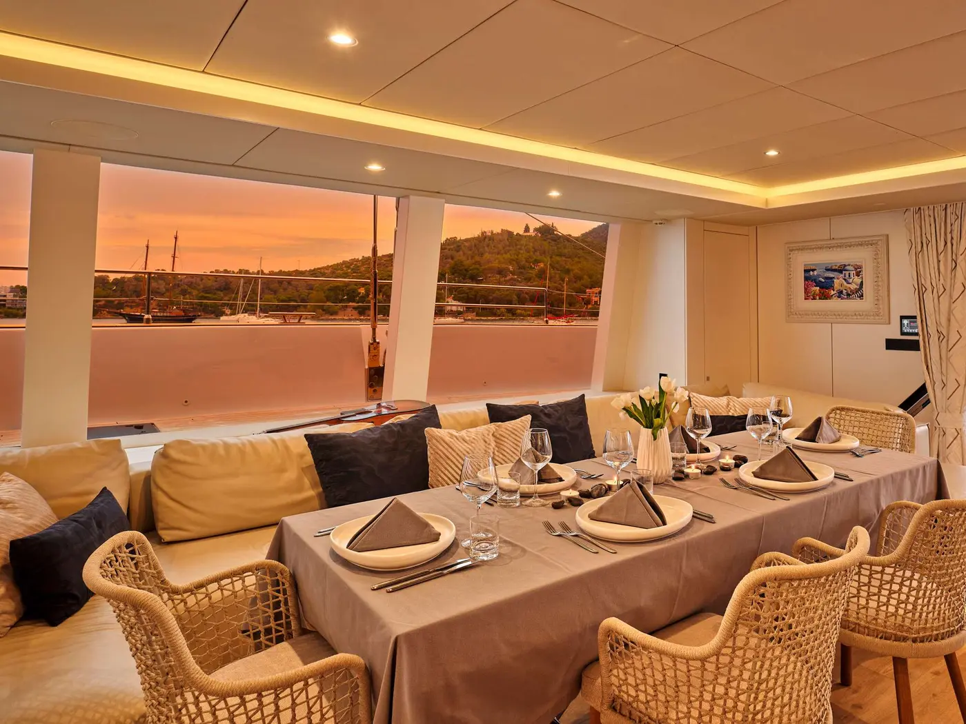 The dining table set for dinner in the saloon of sailing catamaran Genny, with evening light over the harbour beyond