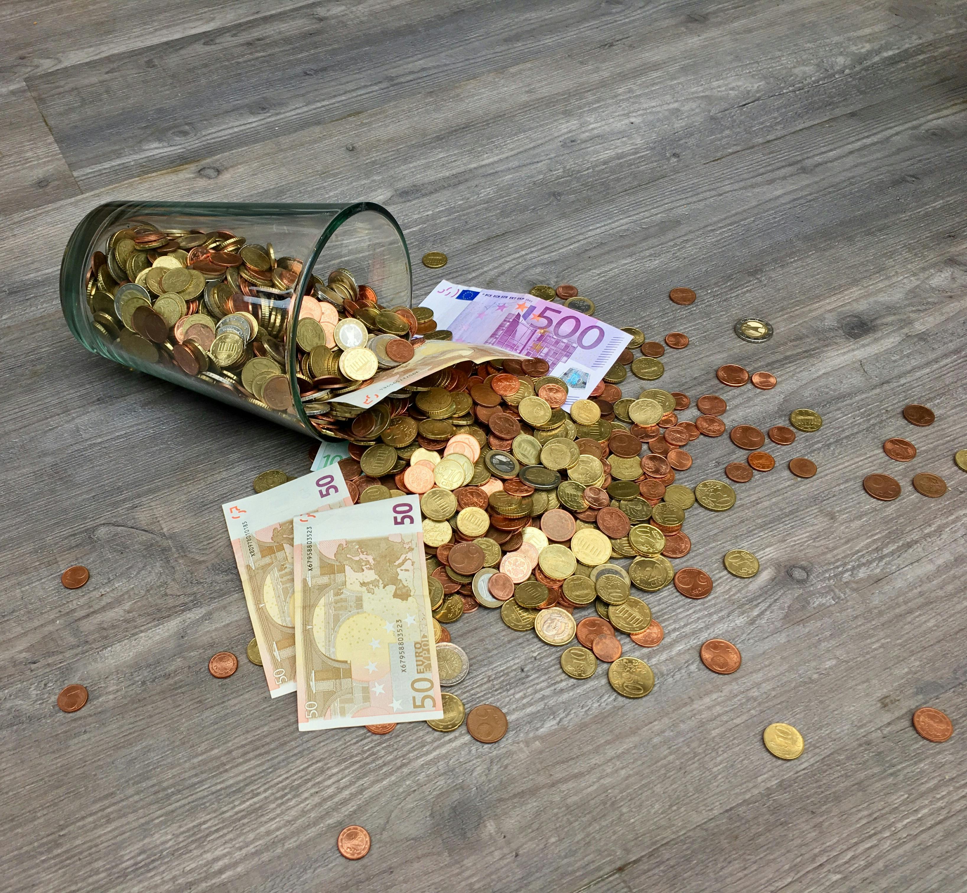 Euro banknotes and coins spilling from a glass jar on a wooden floor, symbolizing expensive yacht charter mistakes and hidden costs in Greece.