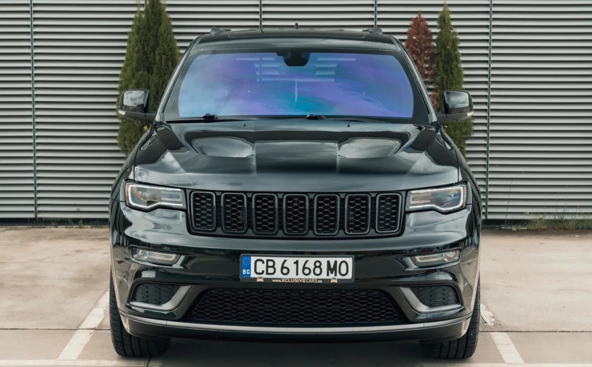 Черен джип Jeep Grand Cherokee, изглед отпред, паркиран пред сива метална стена.