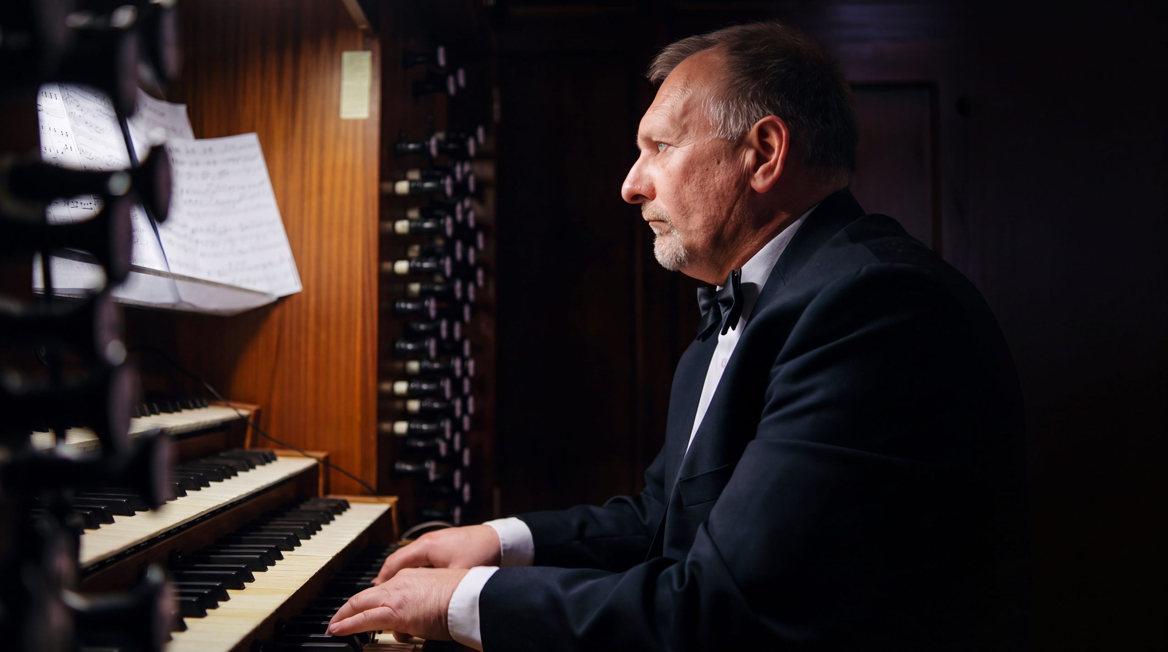 Jan Kępiński we fraku i muszce, z profilu, gra przy kontuarze organów w półmroku
