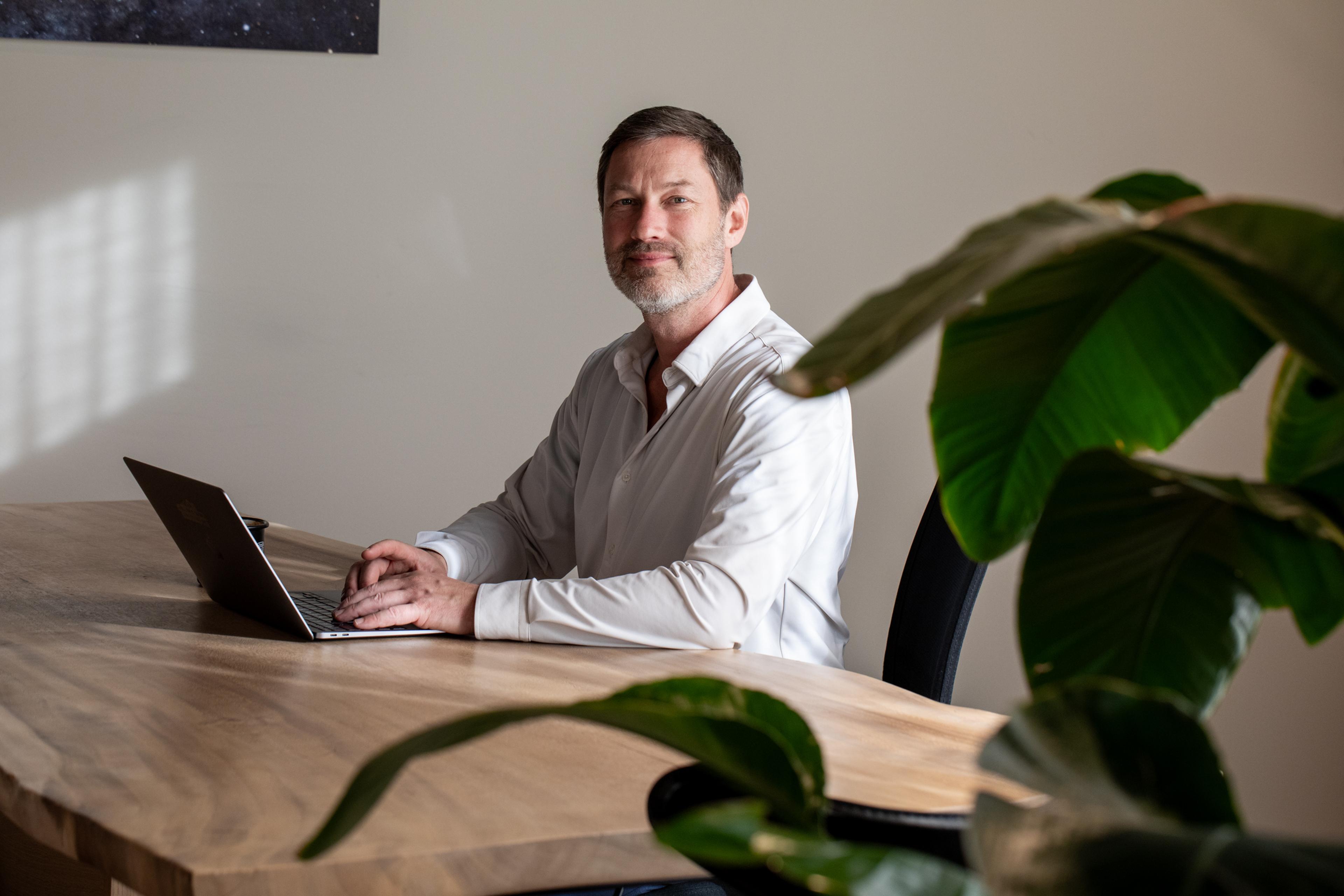 Dr. Lowan Stewart seated at a wooden desk with an open laptop, dressed in a white shirt