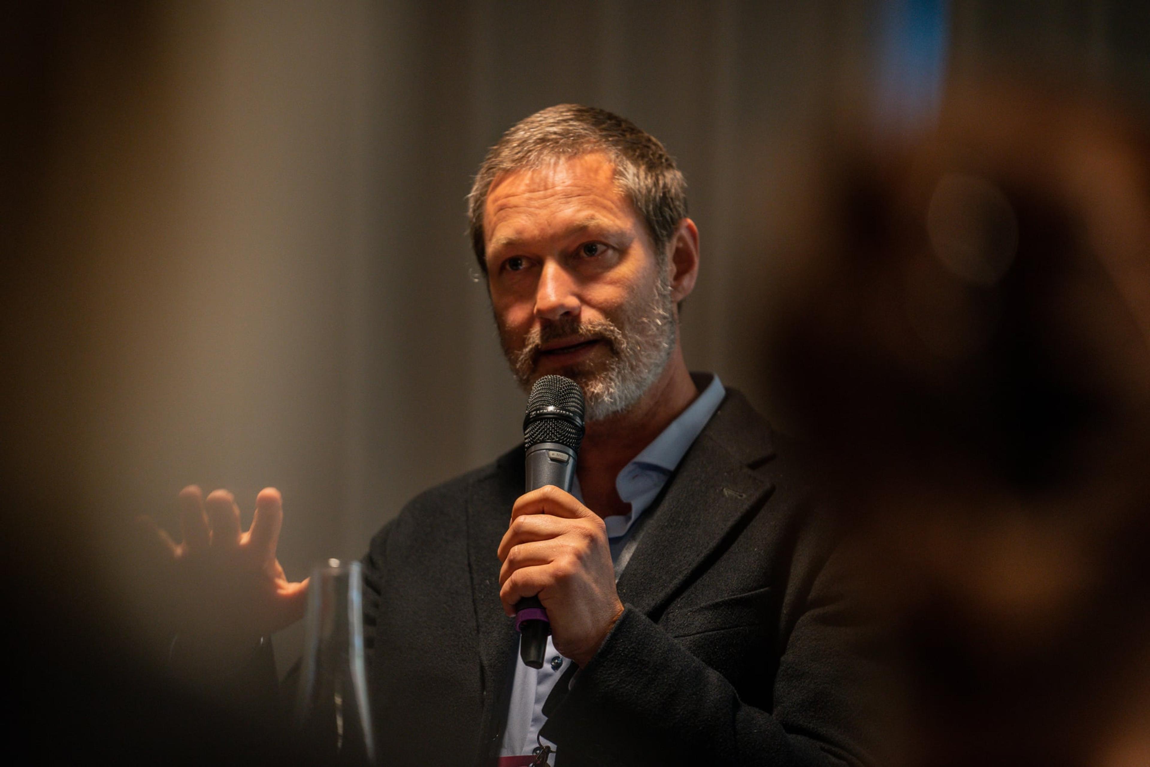 Dr. Lowan Stewart speaking at a podium, holding a microphone, warm stage lighting