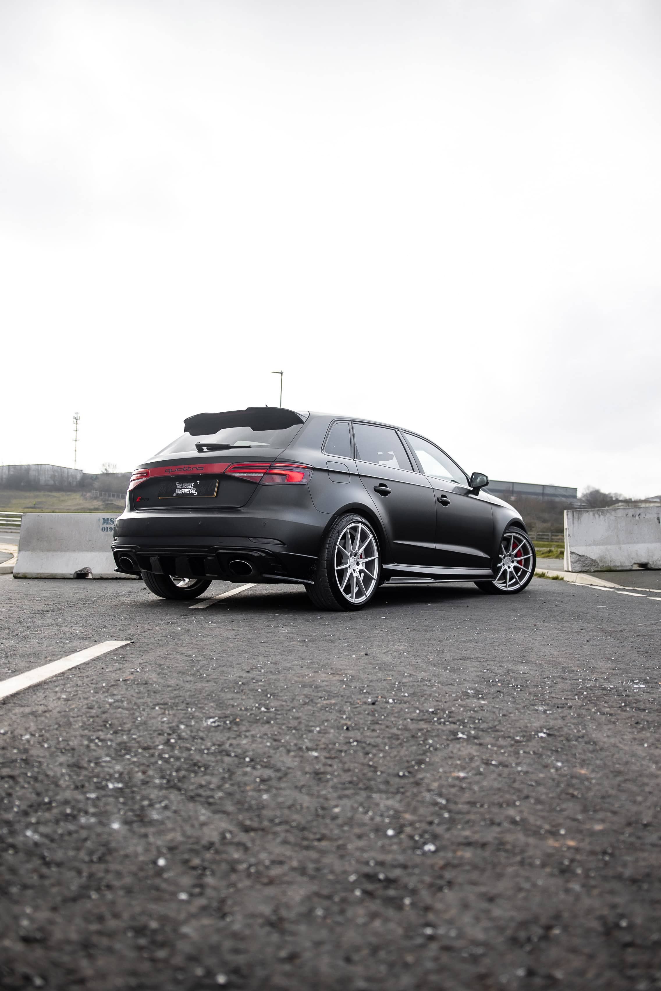 A custom Forge Audi RS3 in matte black sits parked on an asphalt lot between concrete barriers under an overcast sky.