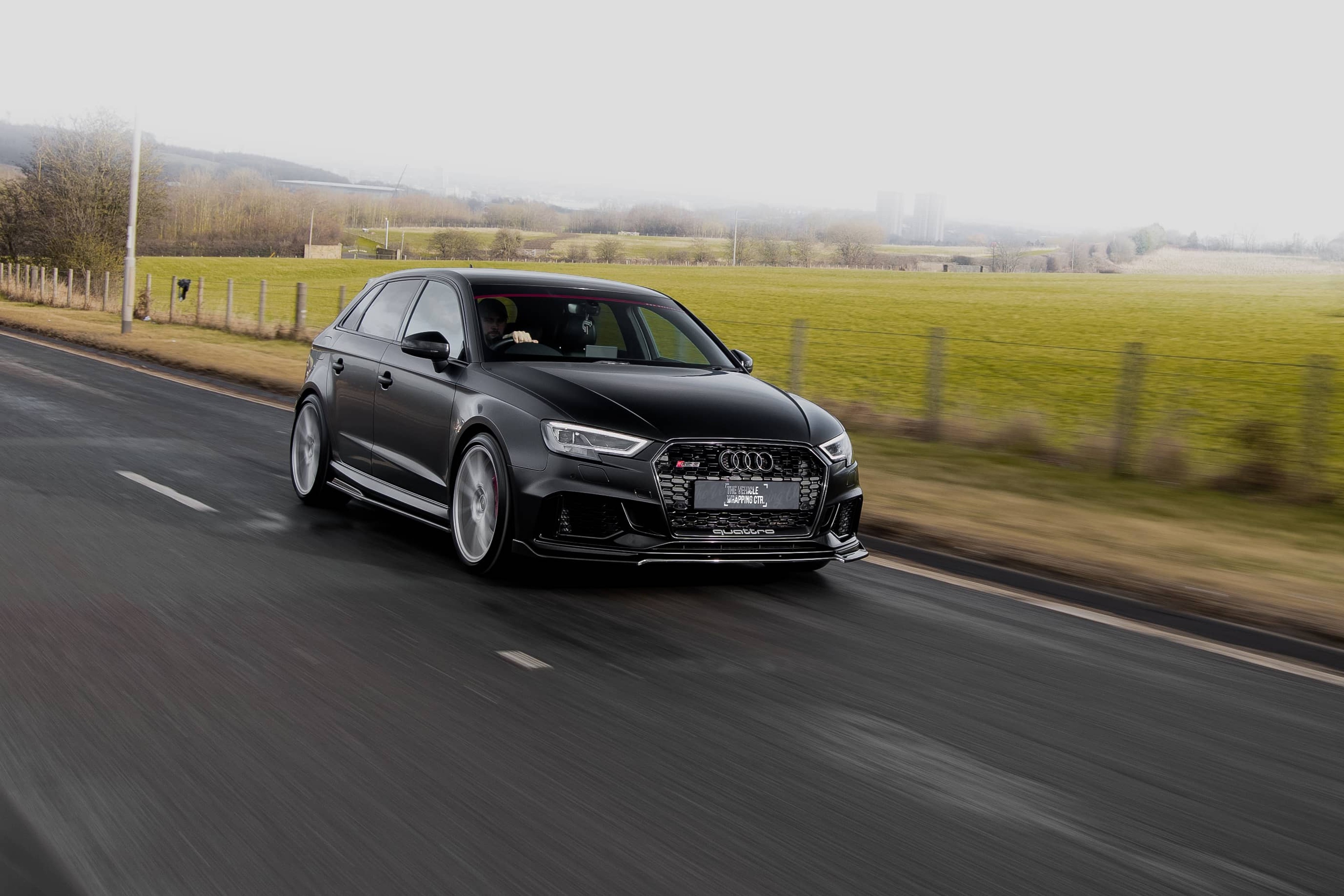 A matte black custom Forge Audi RS3 drives along an open asphalt road alongside a grassy field.