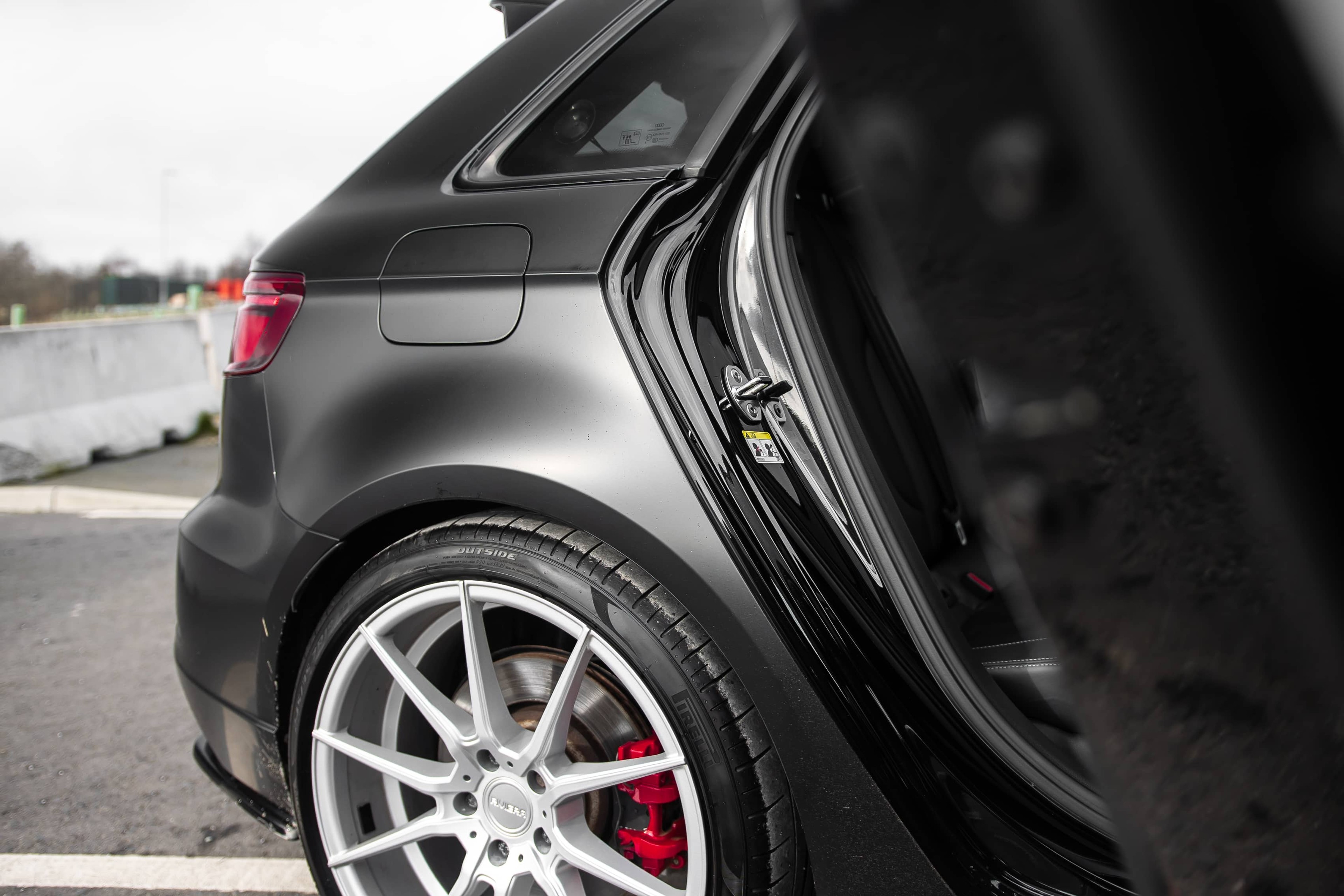 The open rear door of a matte black custom Forge Audi RS3 reveals its sleek wheel arch and silver alloy wheel outdoors.