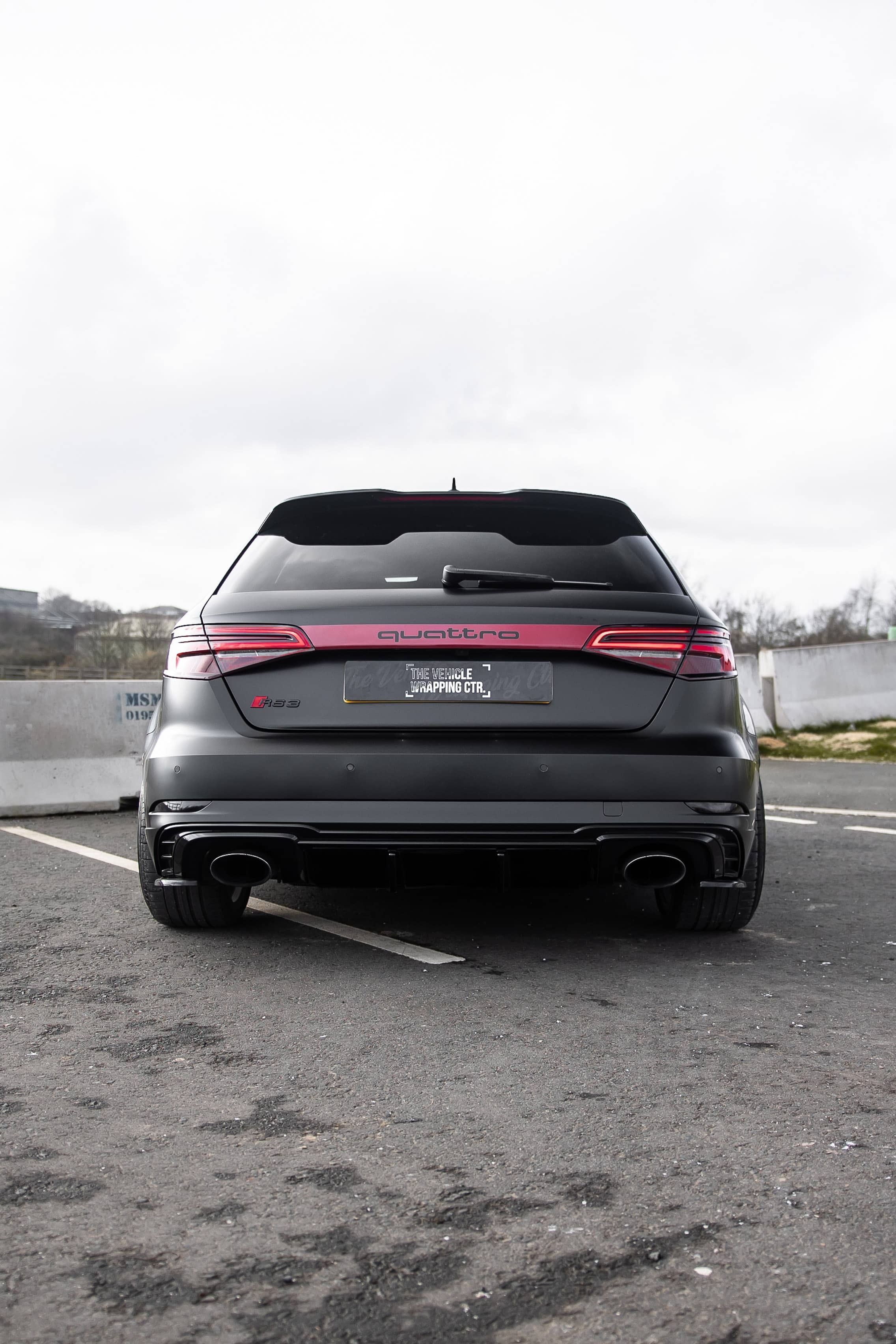 The rear view of a custom Forge Audi RS3 in matte black features glowing taillights and a red quattro strip while parked on asphalt under an overcast sky.