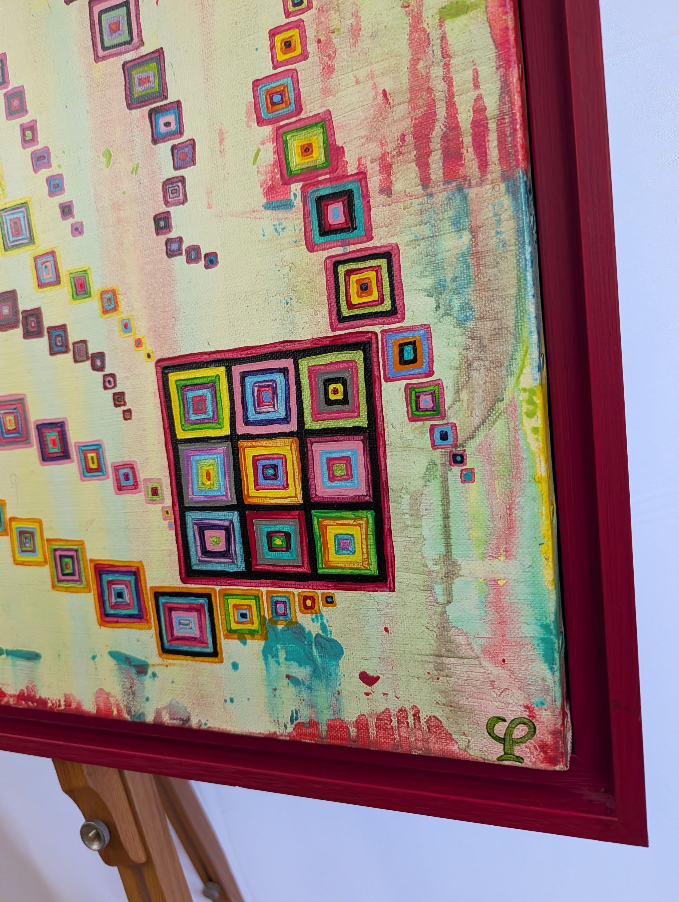 Détail lumineux "Pixels en fête" de Caroline Pons. Carrés concentriques multicolores en pointillisme sur fond clair. Une toile acrylique contemporaine, encadrée de rouge sur chevalet.