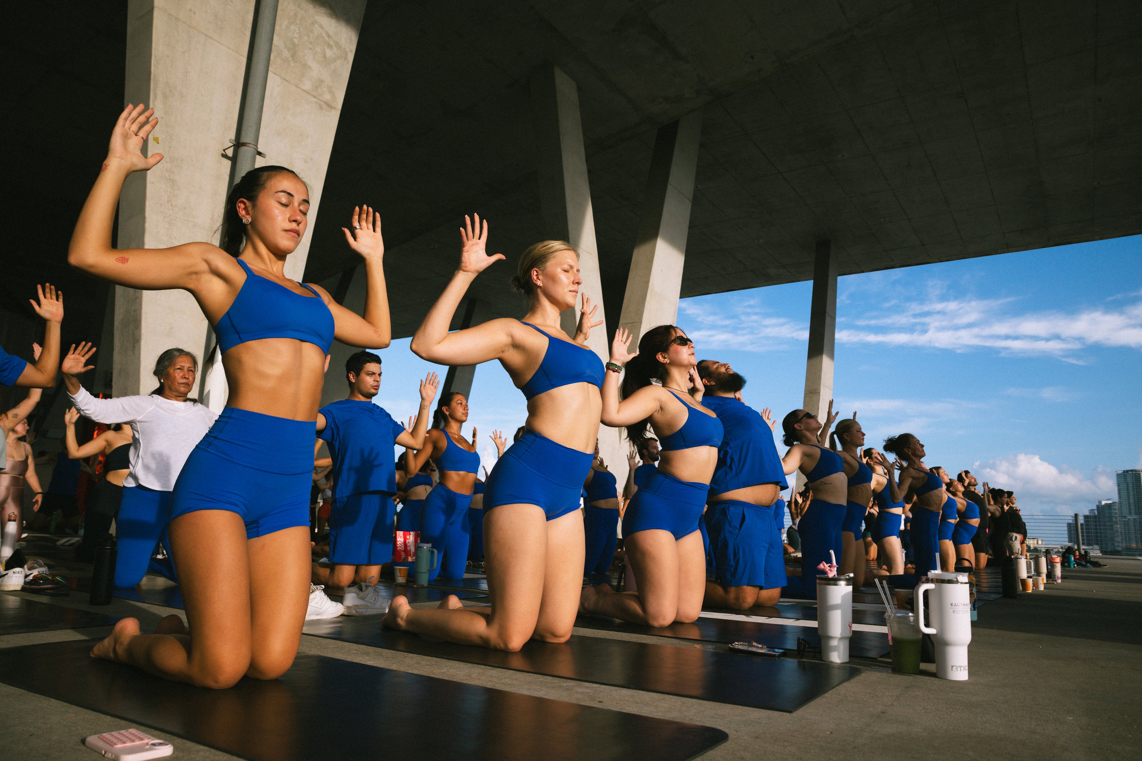 Participants in blue activewear kneel on black mats with their arms raised inside the open-sided garage.