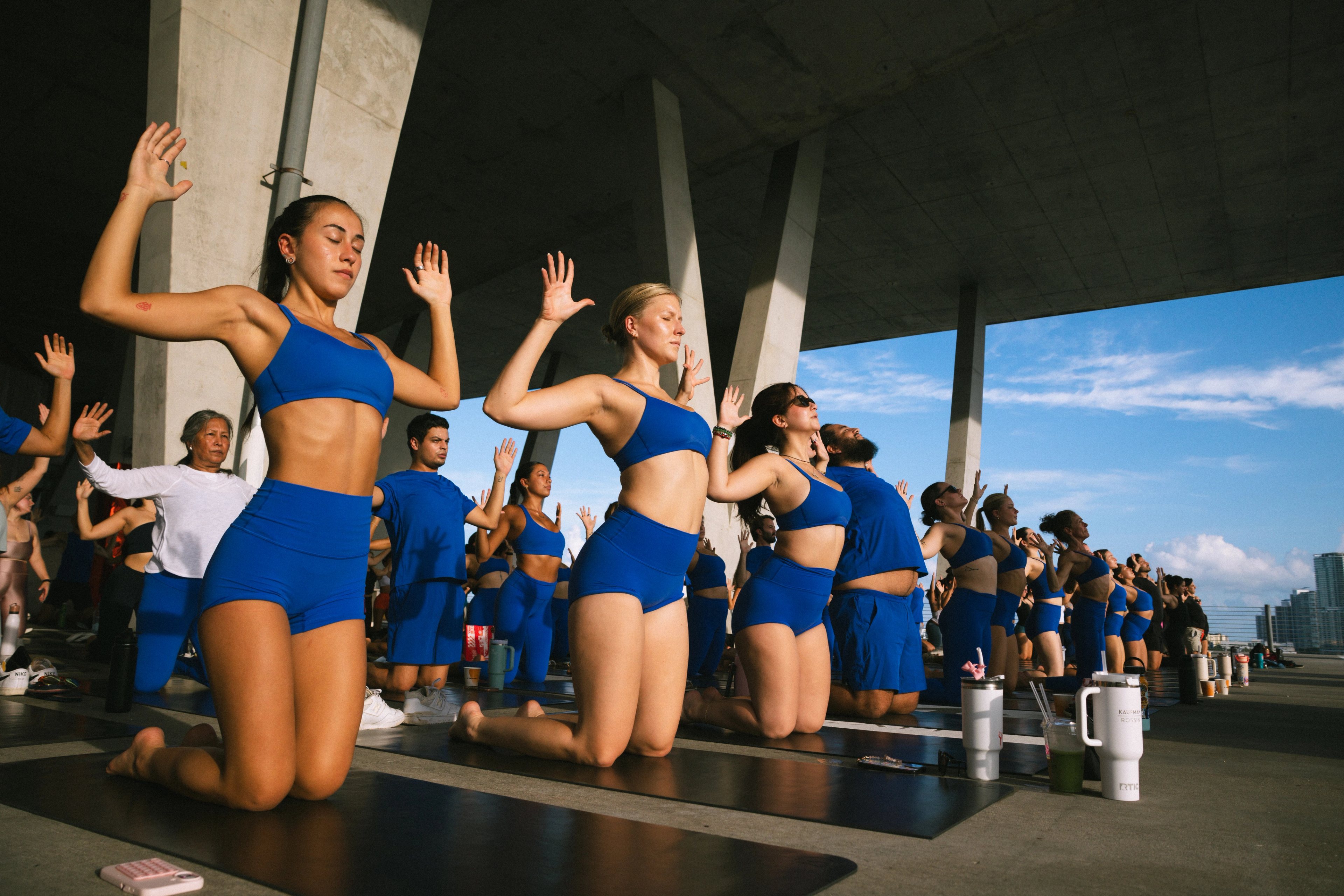 Participants in blue activewear kneel on black mats with their arms raised inside the open-sided garage.