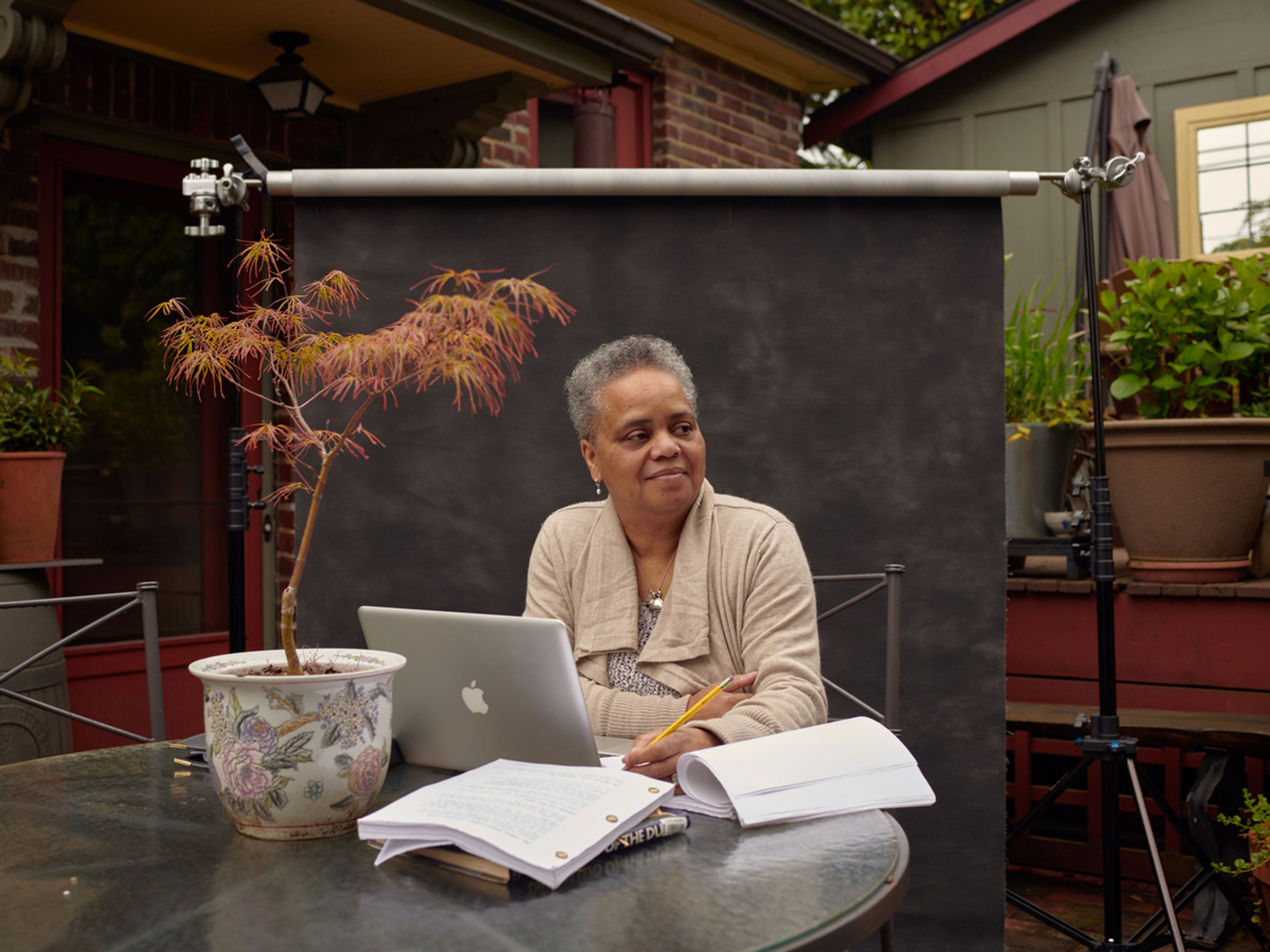 Person sits in front of a black screen in a house, at a round dark table featuring a plant, laptop and various papers. She is wearing a beige jacket and her hair is gray and short.
