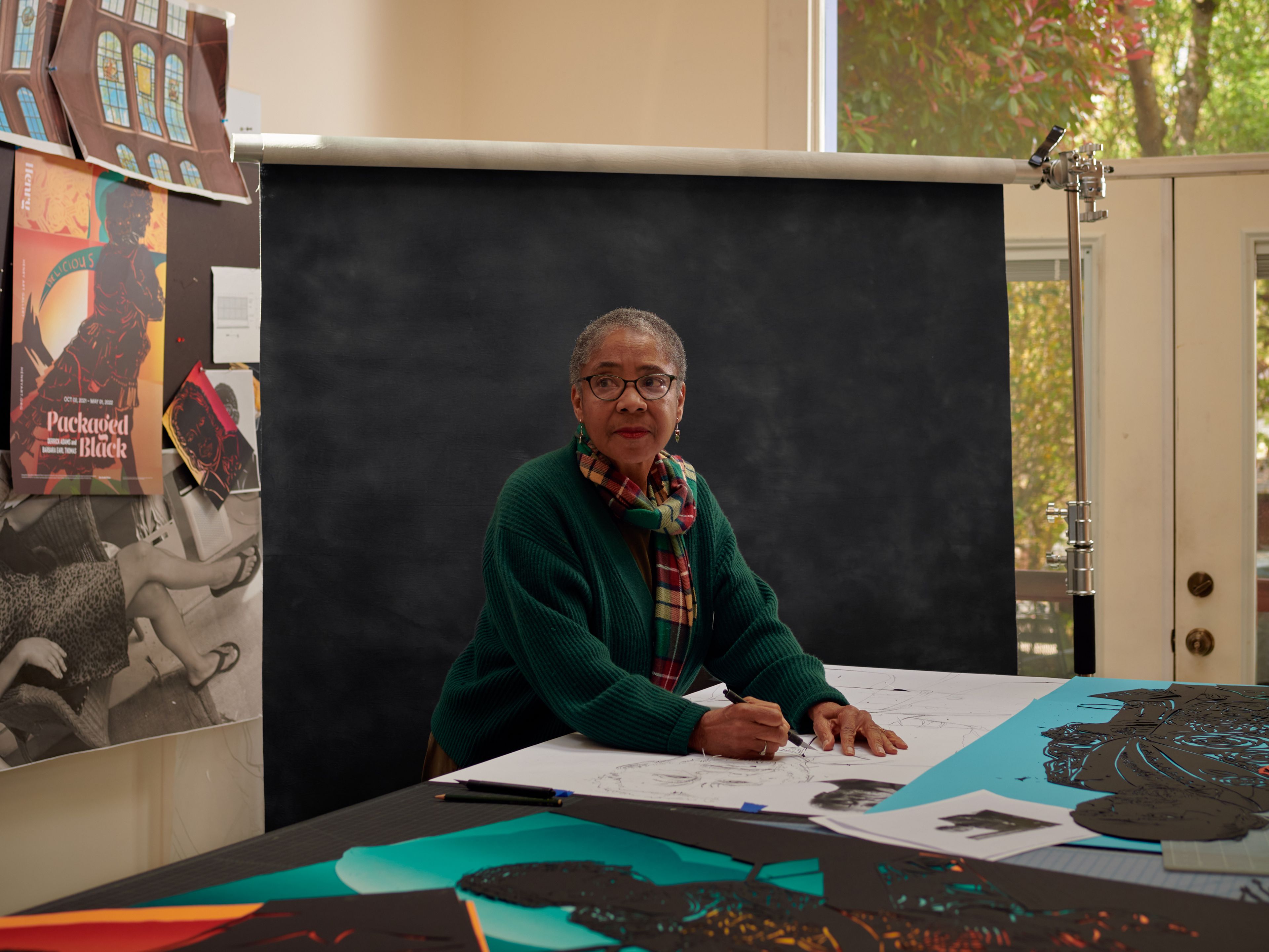 Person in front of a black backdrop in a room with a window to the right. In front of her is a table featuring paper on which she is drawing. It also features colorful papercut artwork.