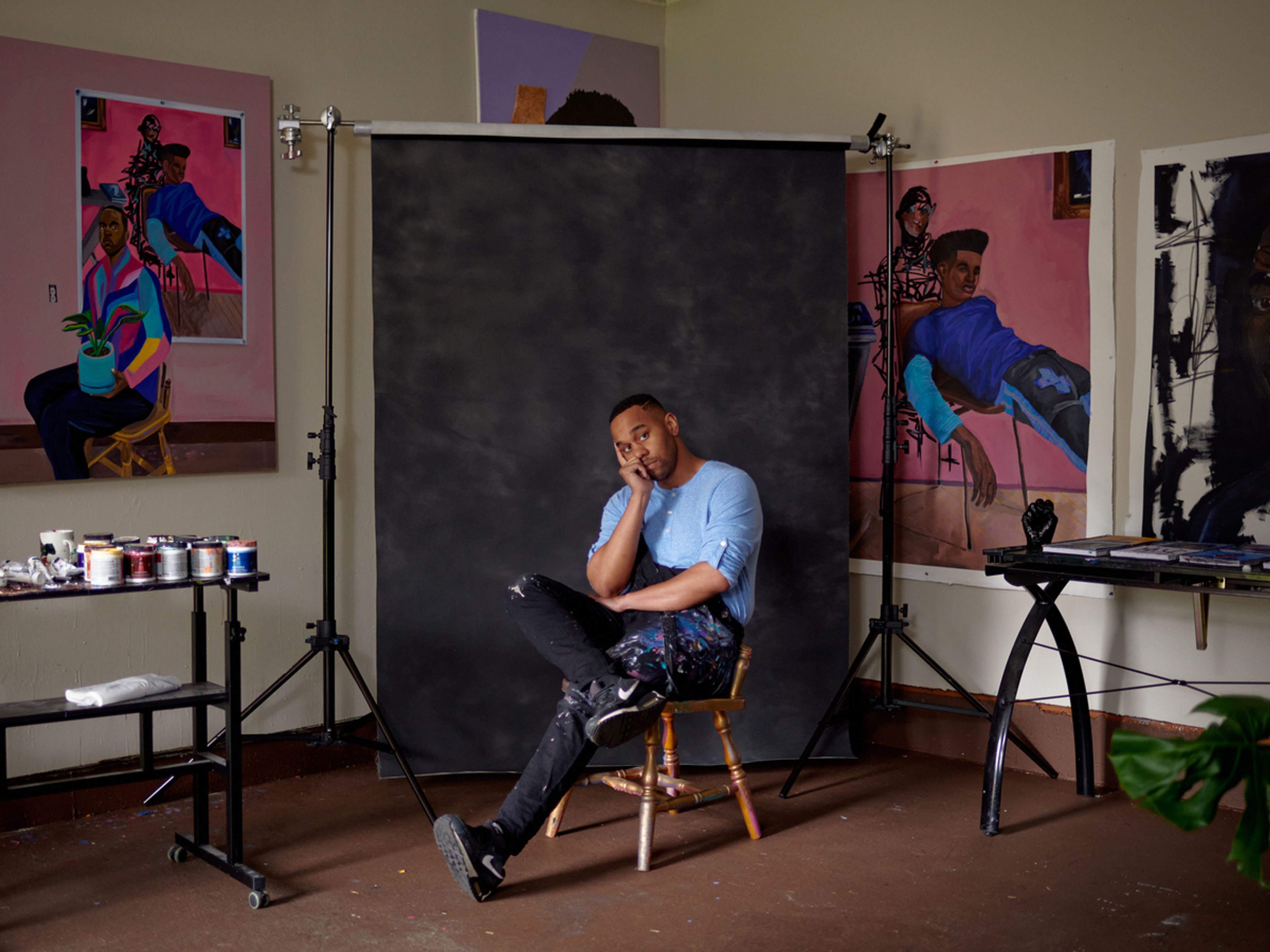 Person on chair in front of black screen, paintings in pink and other bright colors hang on the wall