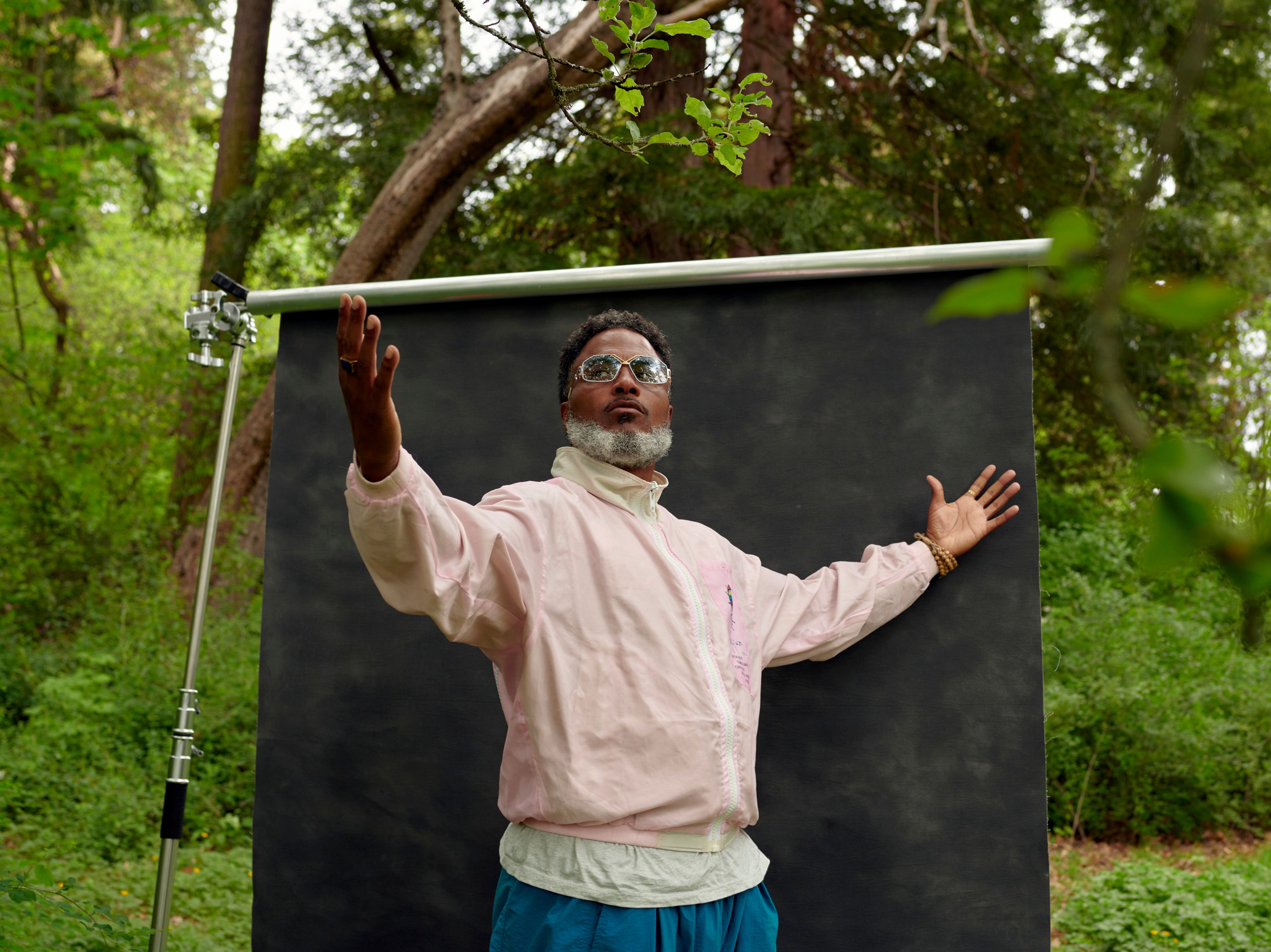 A man wearing a pink jacket and sunglasses stands with arms outstretched in front of a gray back drop surrounded by greenery